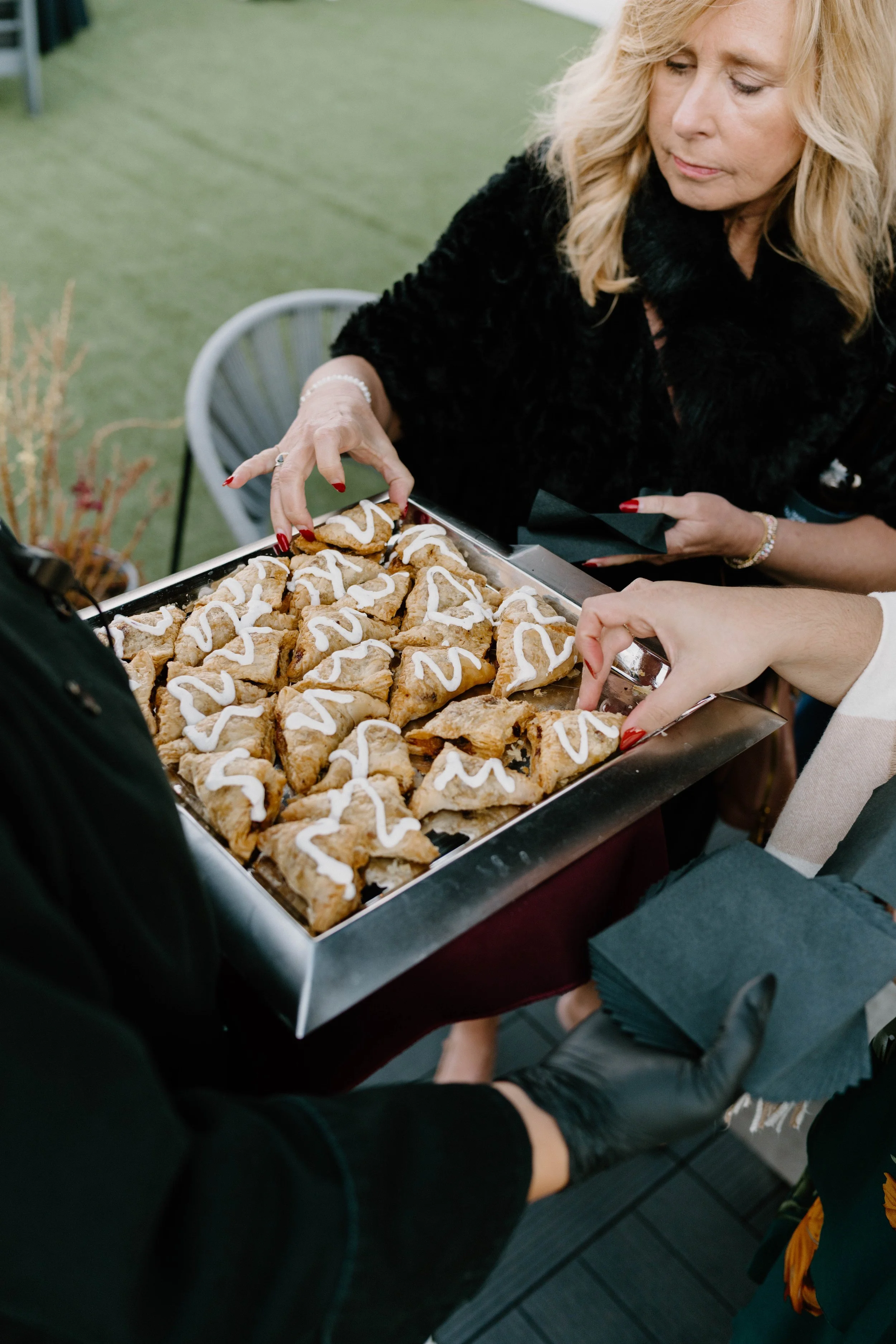 A woman with blonde hair and red nail polish reaching for a tray of Christmas cookies decorated with white icing, while another person is holding the tray with black-gloved hands. The setting appears to be a social gathering or celebration.