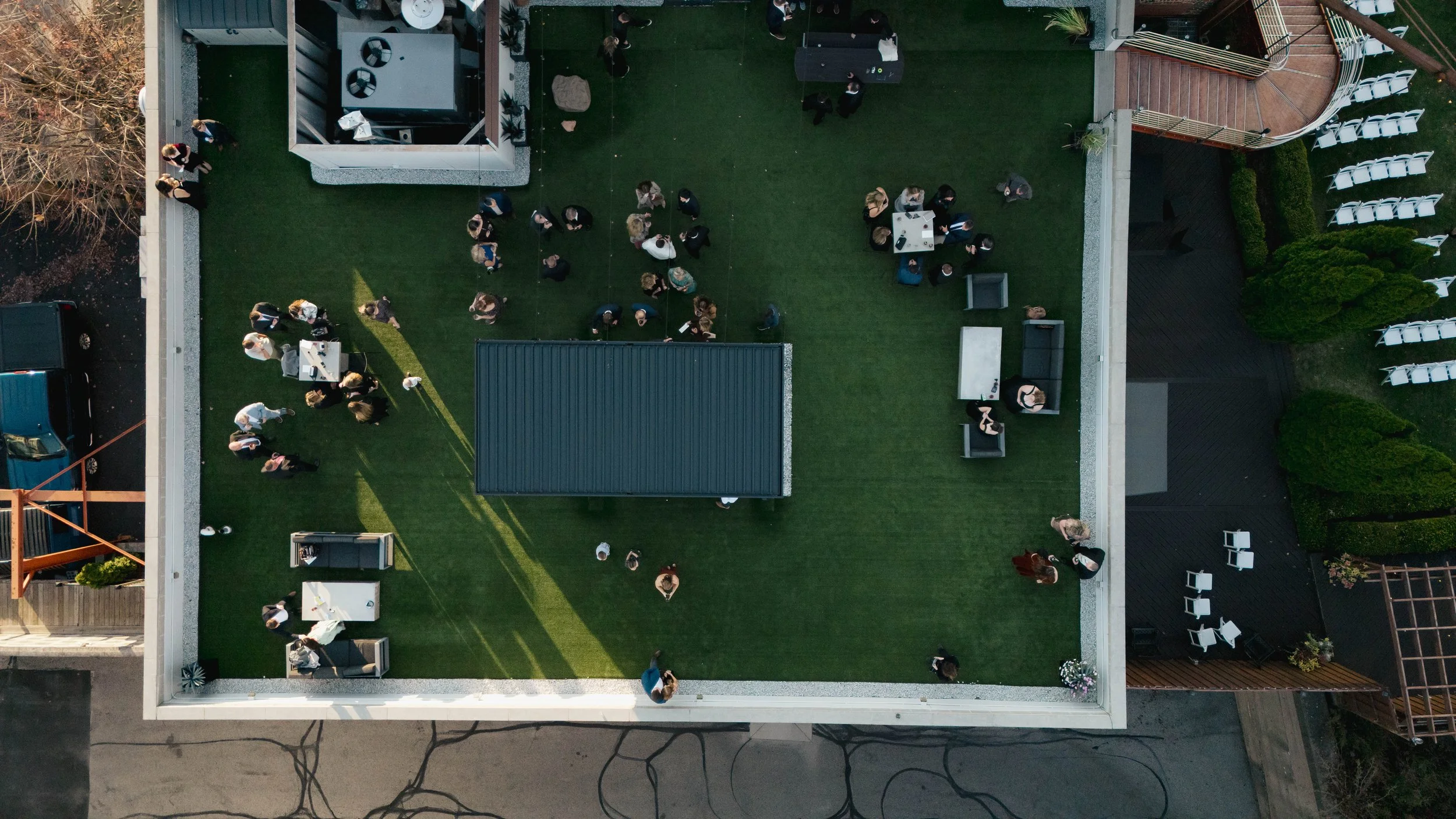 An aerial view of a rooftop patio with people mingling, some standing and some sitting at tables, on a green artificial grass surface. There are outdoor chairs, tables, and plants around the perimeter.