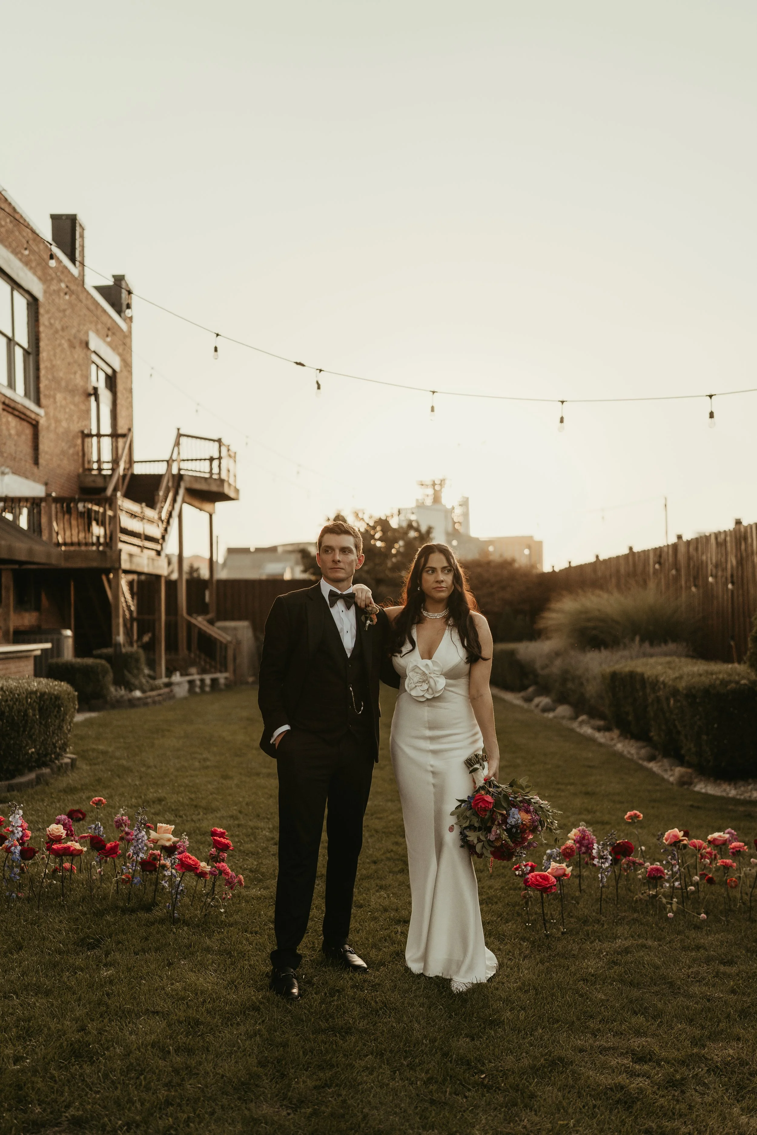 A man in a tuxedo and a woman in a white gown walking arm-in-arm through a garden with colorful flowers, with a house and string lights overhead at sunset.
