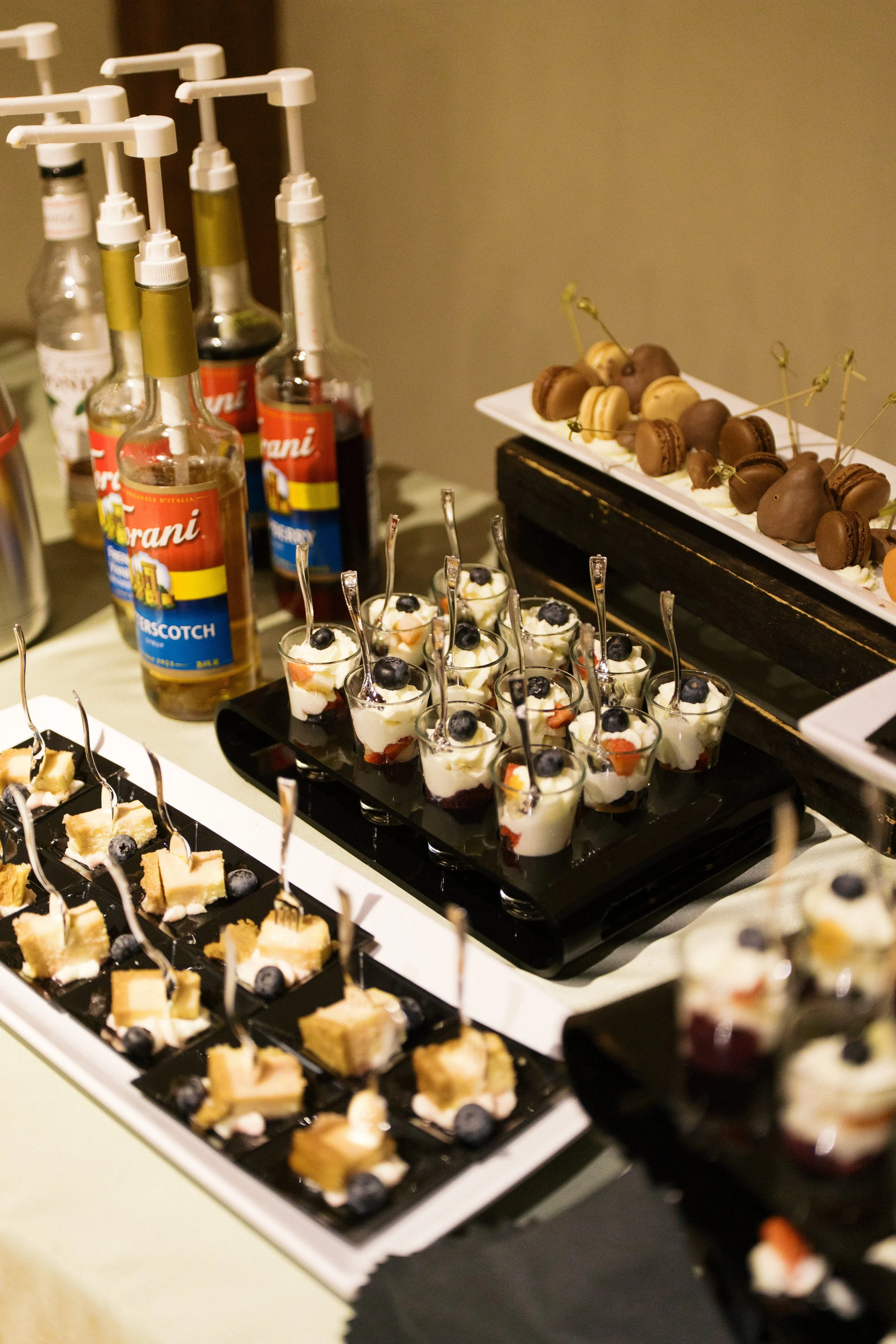 Assorted desserts including small glasses of cream topped with blueberries and other small treats, along with chocolate macarons and other confections on display.