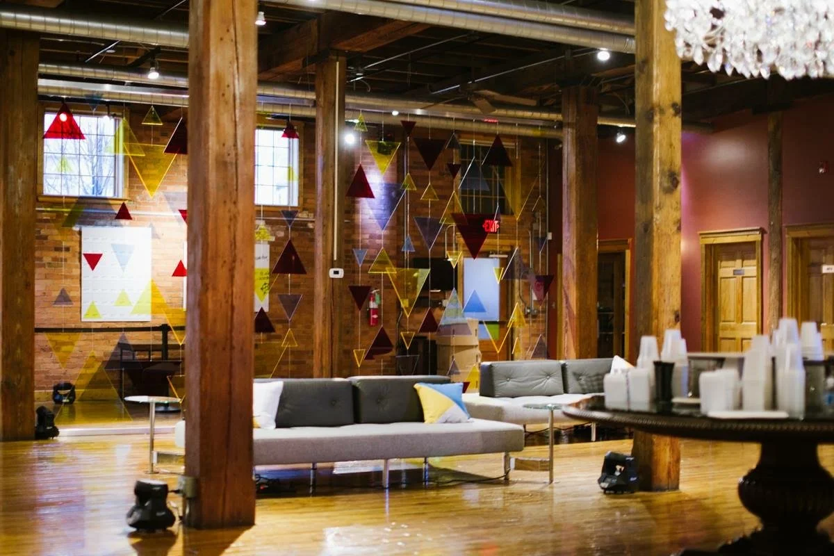 Indoor space with wooden beams and walls, decorated with colorful triangular hanging decorations, large windows letting in natural light, a gray couch with pillows, and a table with cups and bottles.