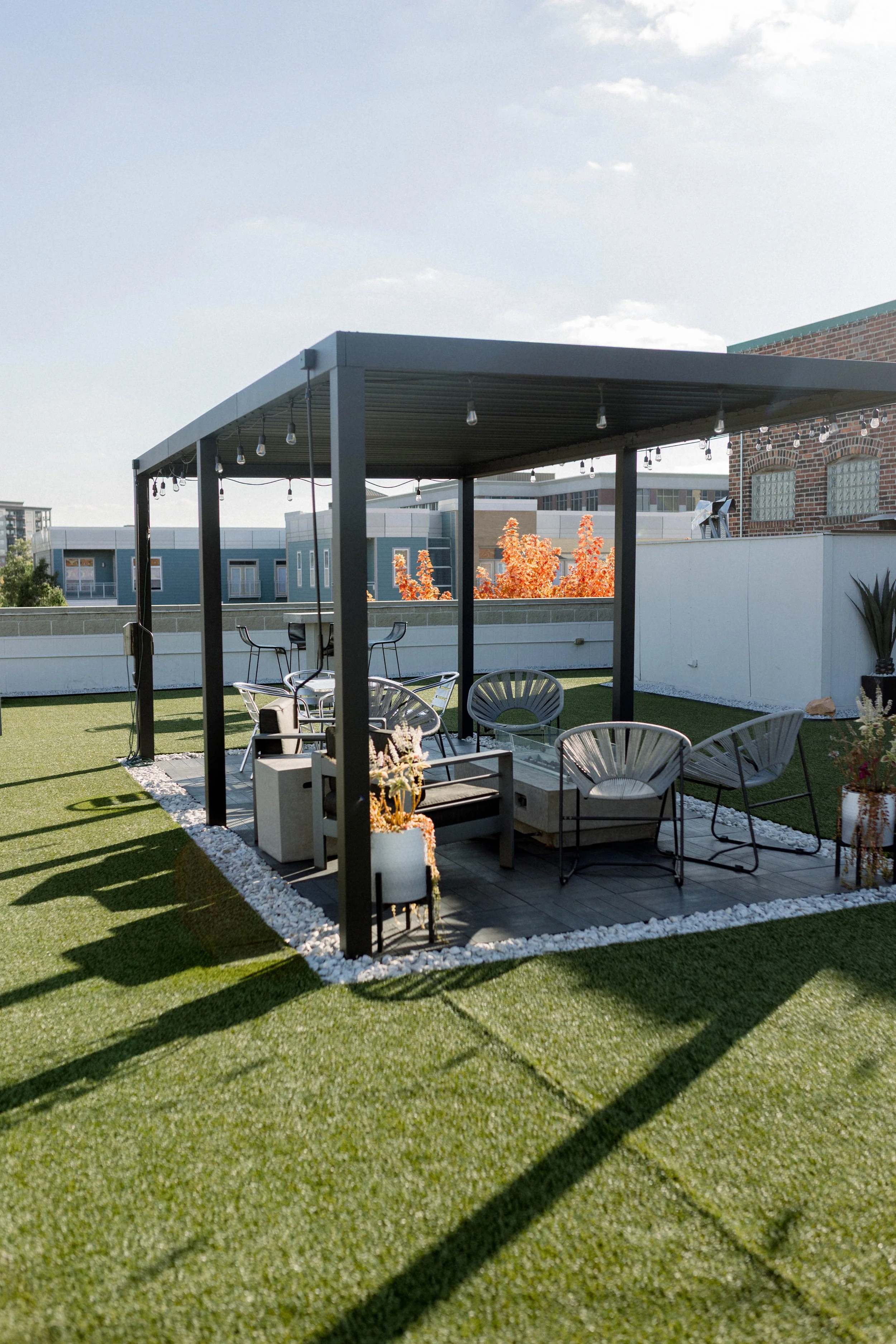A rooftop patio with outdoor furniture, including chairs and a table, under a metal pergola with string lights. The area is bordered by white decorative stones and has artificial green grass. In the background, there are neighboring buildings, trees,