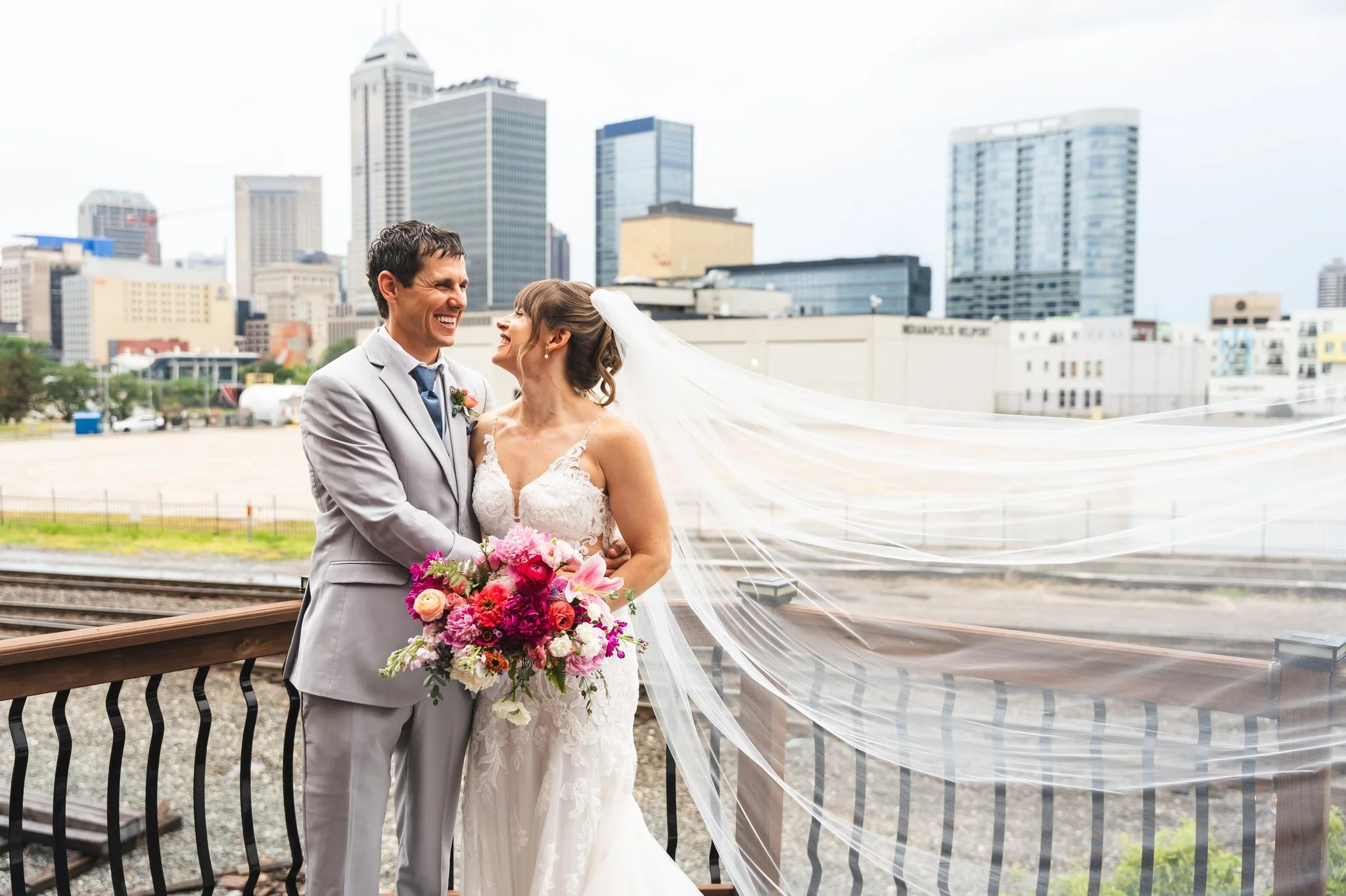 A bride and groom on a train track in an urban area, smiling and looking at each other, with the bride holding a bouquet of vibrant pink, orange, and white flowers, and the groom in a light gray suit with a blue tie, city skyscrapers in the backgroun