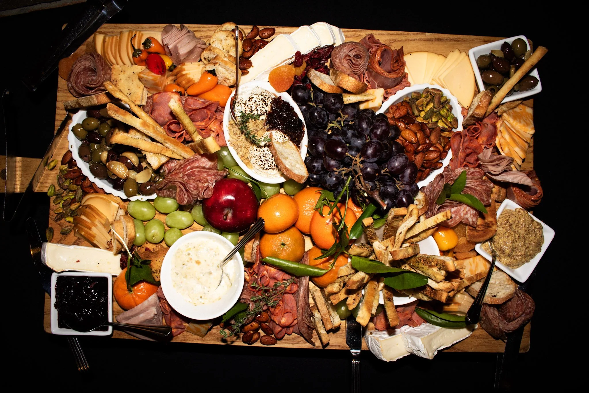 A tray of assorted cheeses, fruits, nuts, bread, meats, and condiments on a wooden platter, including grapes, oranges, apples, almonds, pistachios, dates, and various slices of cheese and cured meats.