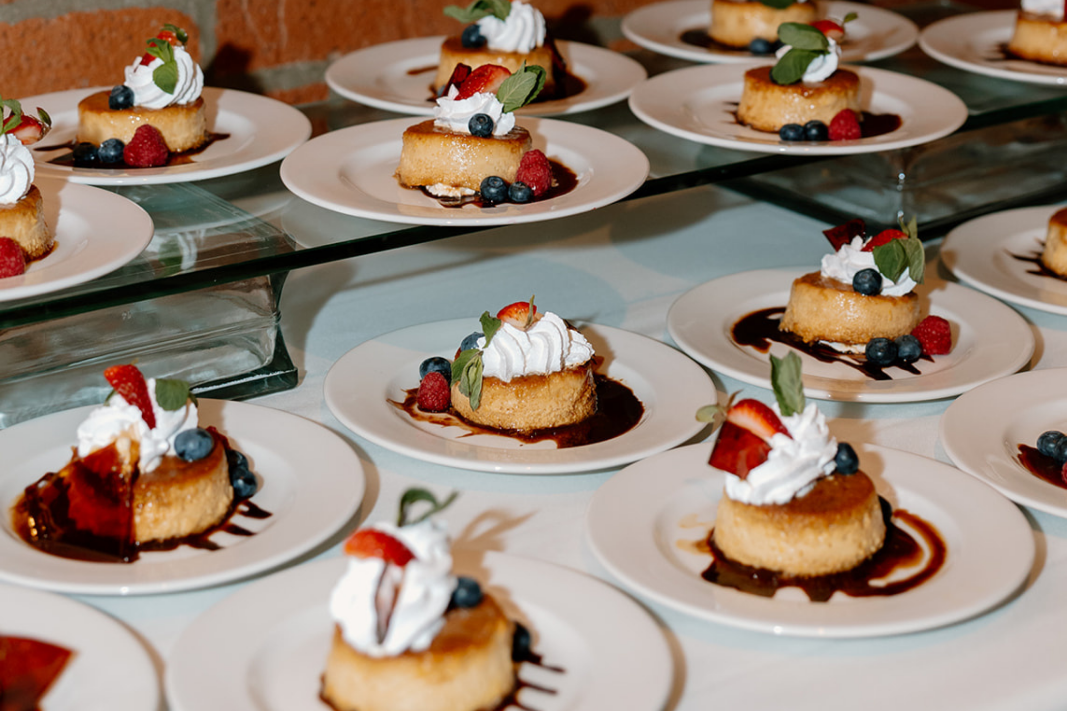 Individual cheesecake slices garnished with whipped cream, strawberries, blueberries, and mint leaves, arranged on white plates with chocolate sauce drizzled underneath, on a glass display stand.