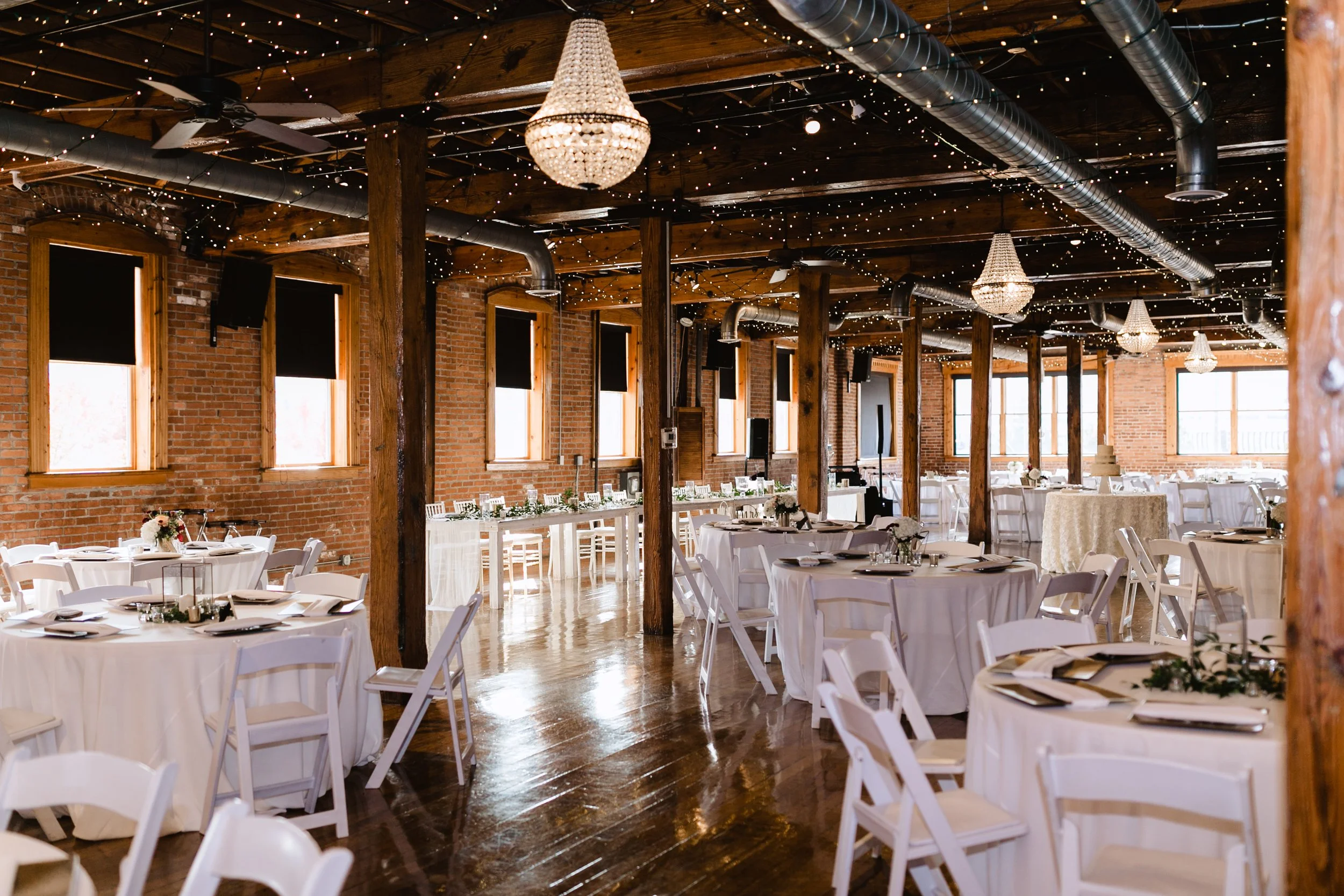 Elegant event space with round tables covered in white tablecloths, decorated with floral centerpieces, set with plates and utensils. The room has exposed brick walls, wooden beams, large windows, and hanging chandeliers with string lights creating a