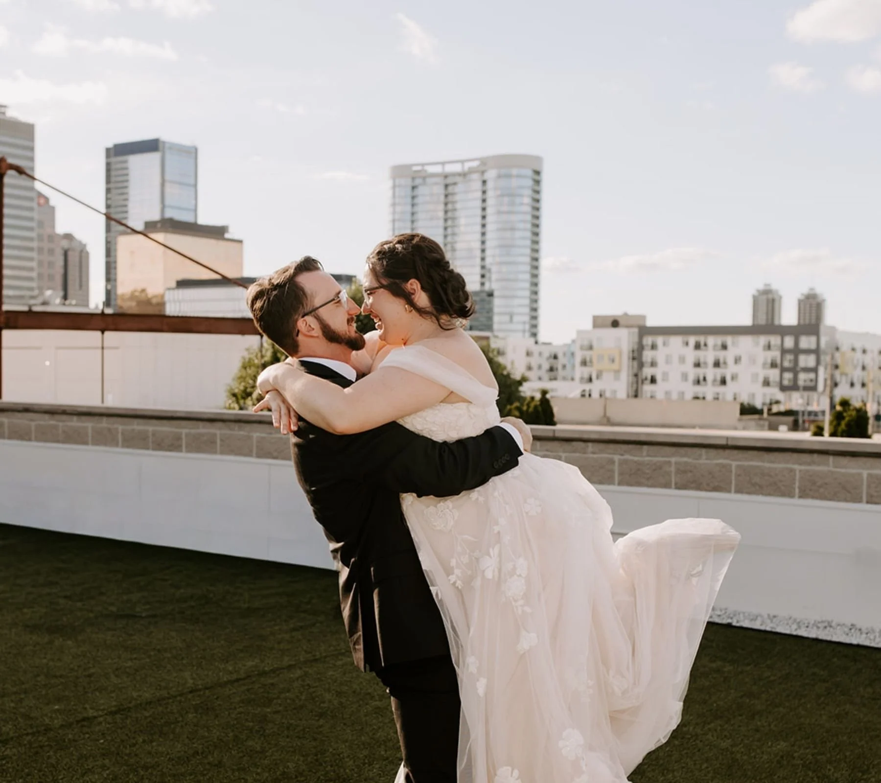 A newlywed couple happily celebrating on a rooftop with city buildings in the background. The groom, wearing a black suit and glasses, lifts the bride, who is in a white wedding dress with lace details, into his arms. They are smiling and looking at 