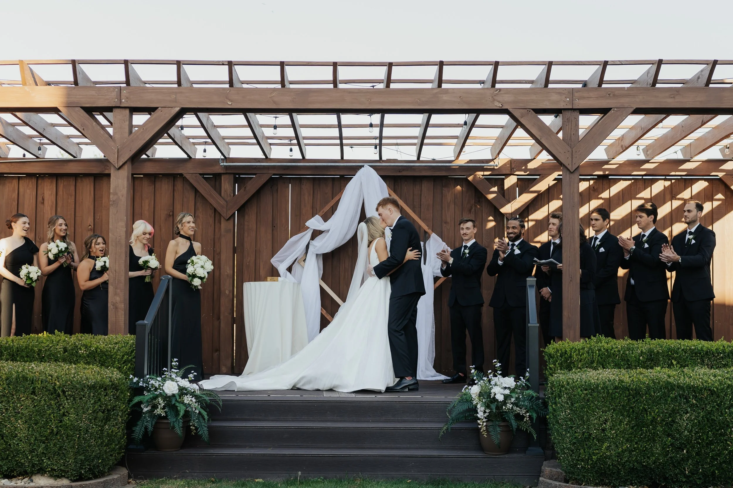 A group of wedding guests watching as a bride and groom kiss during their outdoor ceremony, with bridesmaids holding bouquets on the left and groomsmen standing on the right, under a wooden pergola decorated with a white fabric backdrop and flowers.