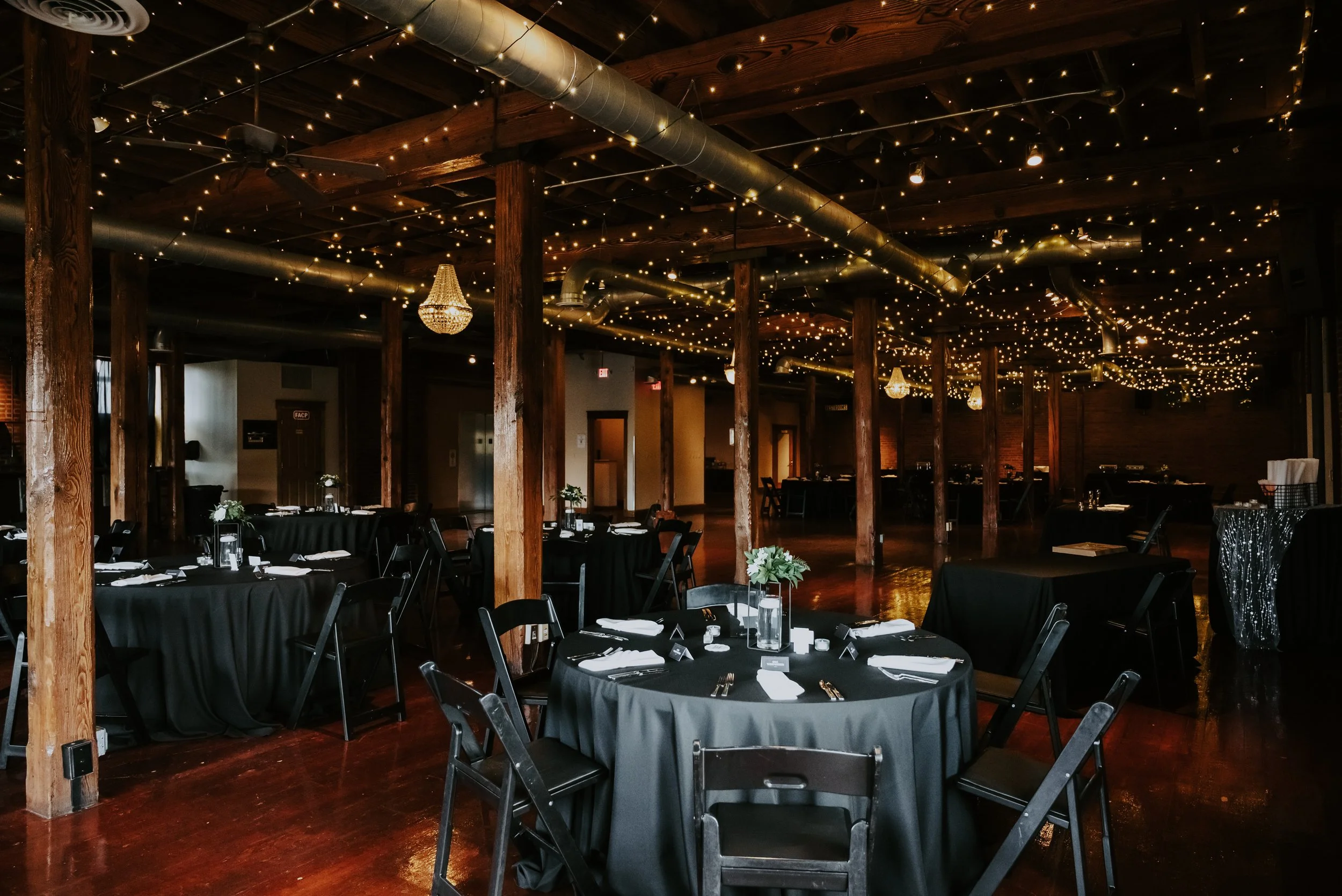 Interior of a decorated event space with black tables, black chairs, and round vases with flowers, illuminated by string lights and chandeliers hanging from the wooden ceiling.