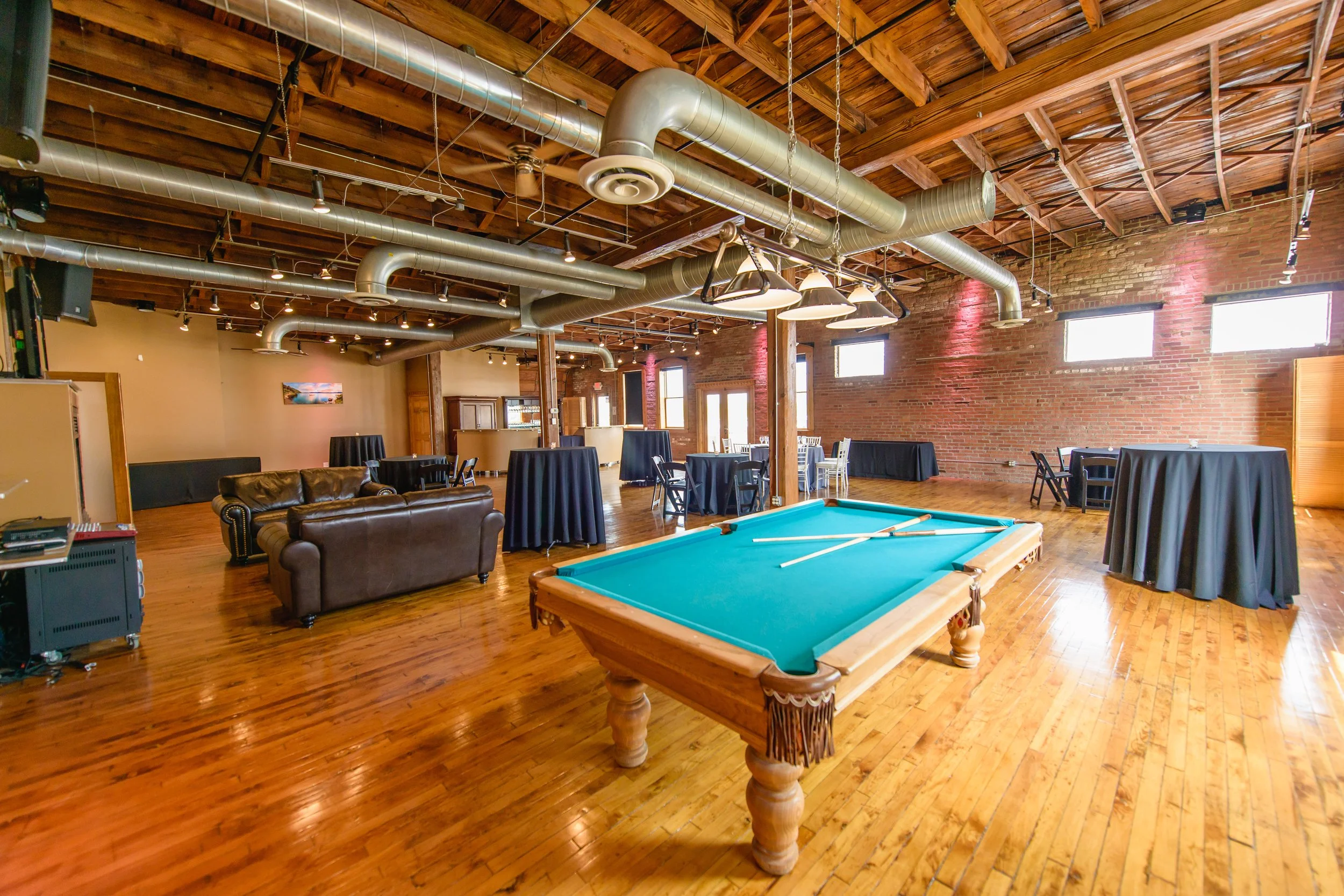A spacious indoor bar or entertainment venue with exposed brick walls, wooden flooring, and a high wooden ceiling with large metallic ventilation pipes. There are several round tables with black tablecloths and chairs, a leather sofa, and a pool tabl