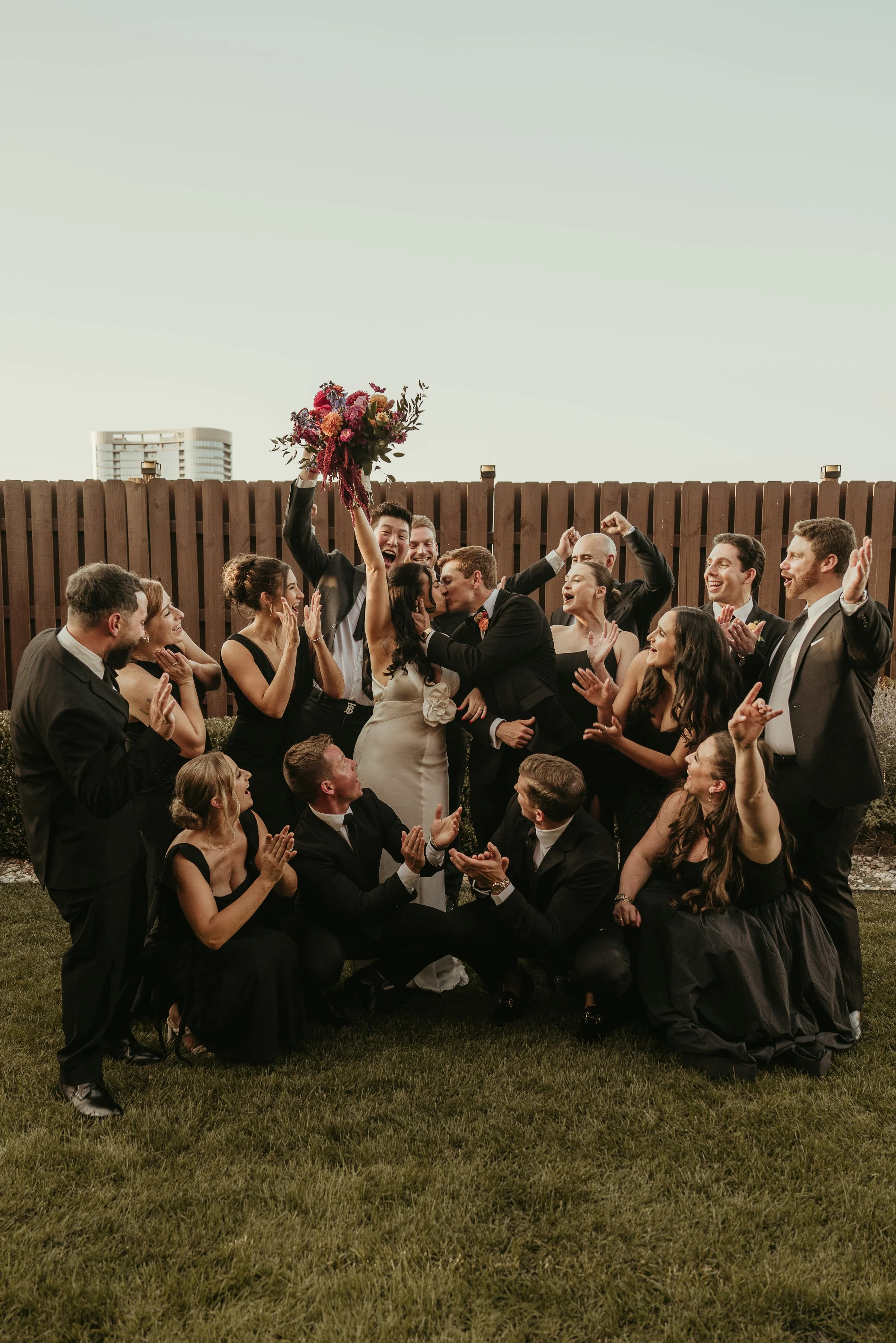 A group of people dressed in formal attire celebrating and cheering outdoors, with a man holding a bouquet of flowers aloft in the center.