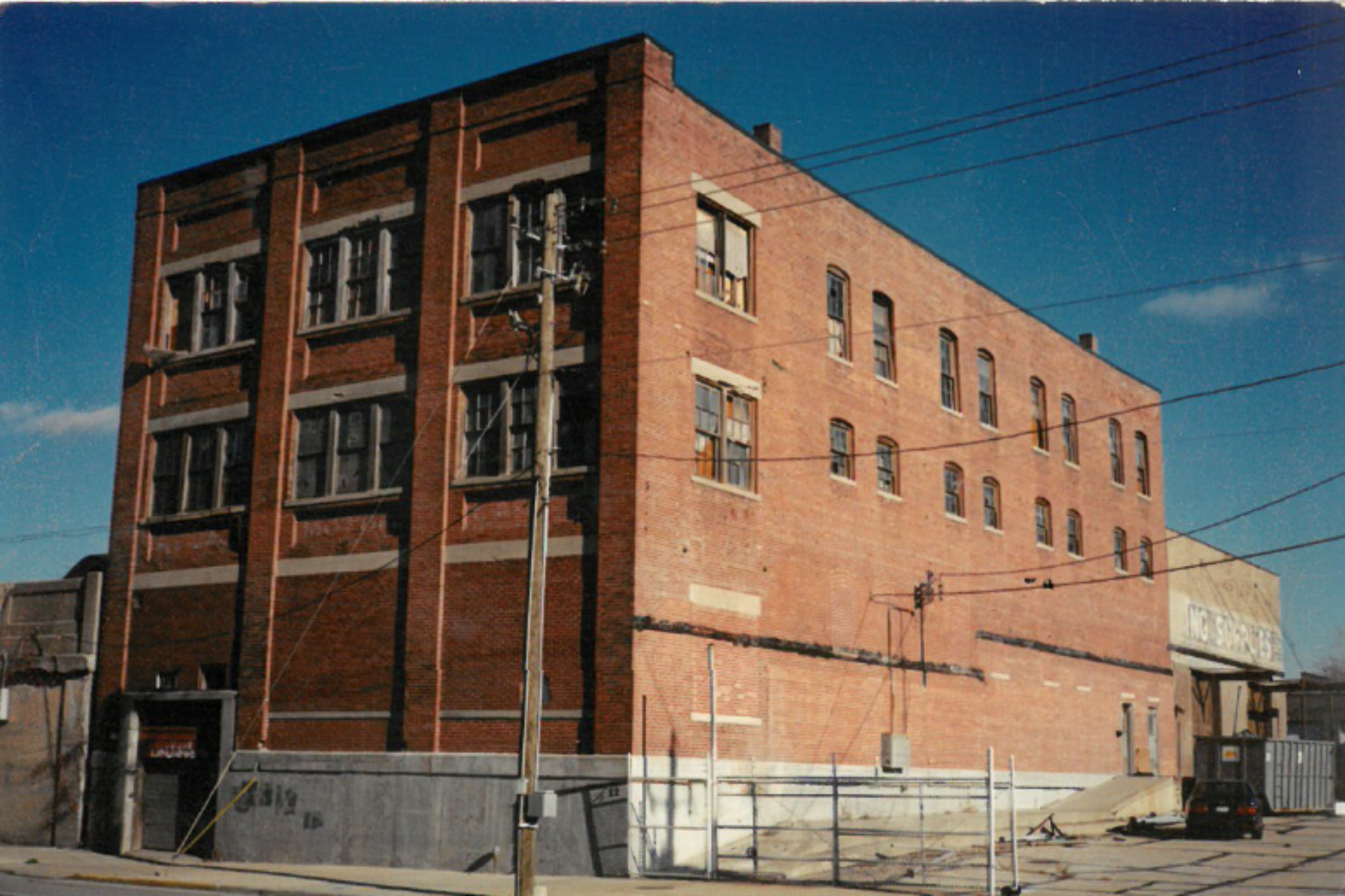 A brick building under construction with several windows and an unfinished ground level. Utility poles with wires are in front of the building, and a clear blue sky is above.