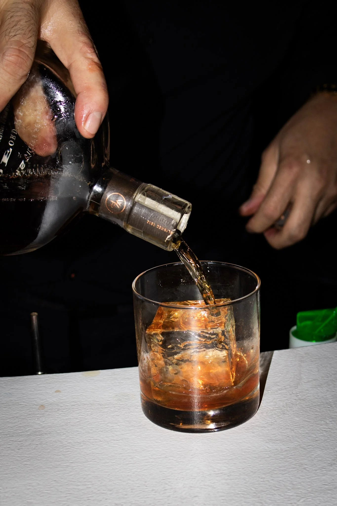 A hand pouring dark liquor from a bottle into a glass filled with ice.