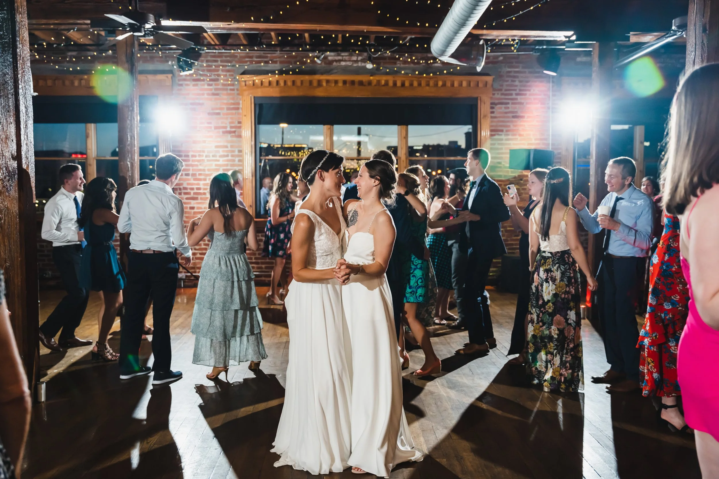 Two women in wedding dresses stand close together, smiling and holding hands on a dance floor. They are surrounded by wedding guests dancing and socializing at an indoor reception, with string lights and brick walls visible in the background.