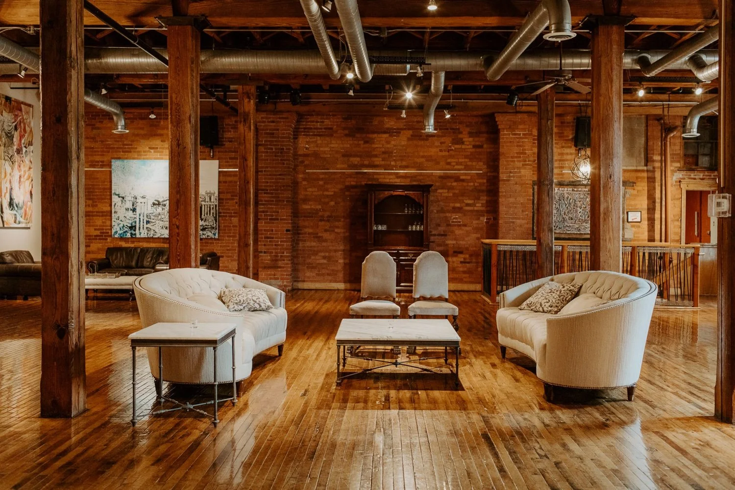 Loft-style interior with exposed brick walls, wooden floor, and ceiling beams. Features two white upholstered sofas with pillows, a coffee table in the center, and two chairs with high backs. Ductwork and lighting fixtures are visible on the ceiling,