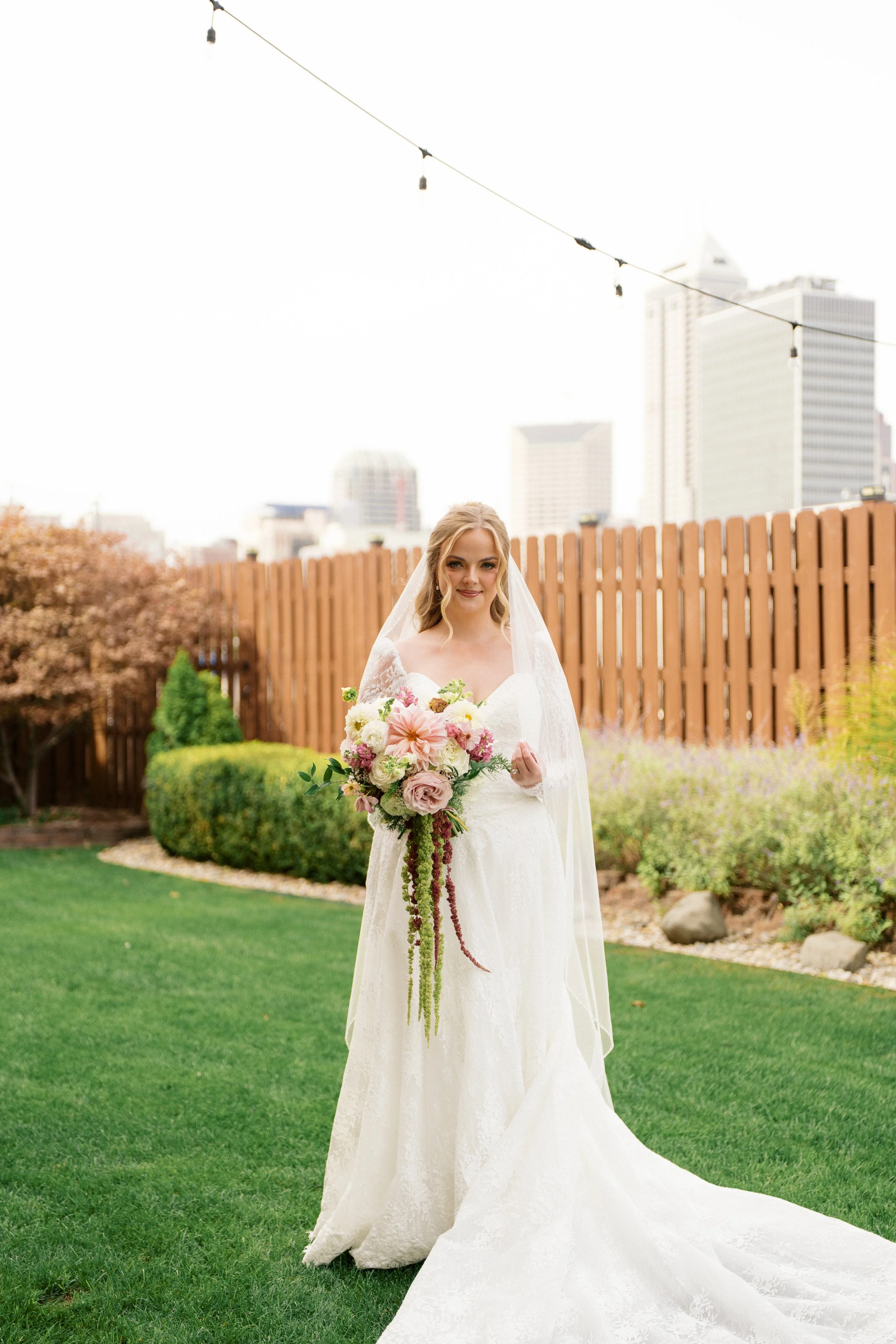 A bride wearing a white wedding dress and veil, holding a large bouquet of pink and white flowers, standing on a well-manicured lawn with trees and a wooden fence in the background, and city skyscrapers in the distance.