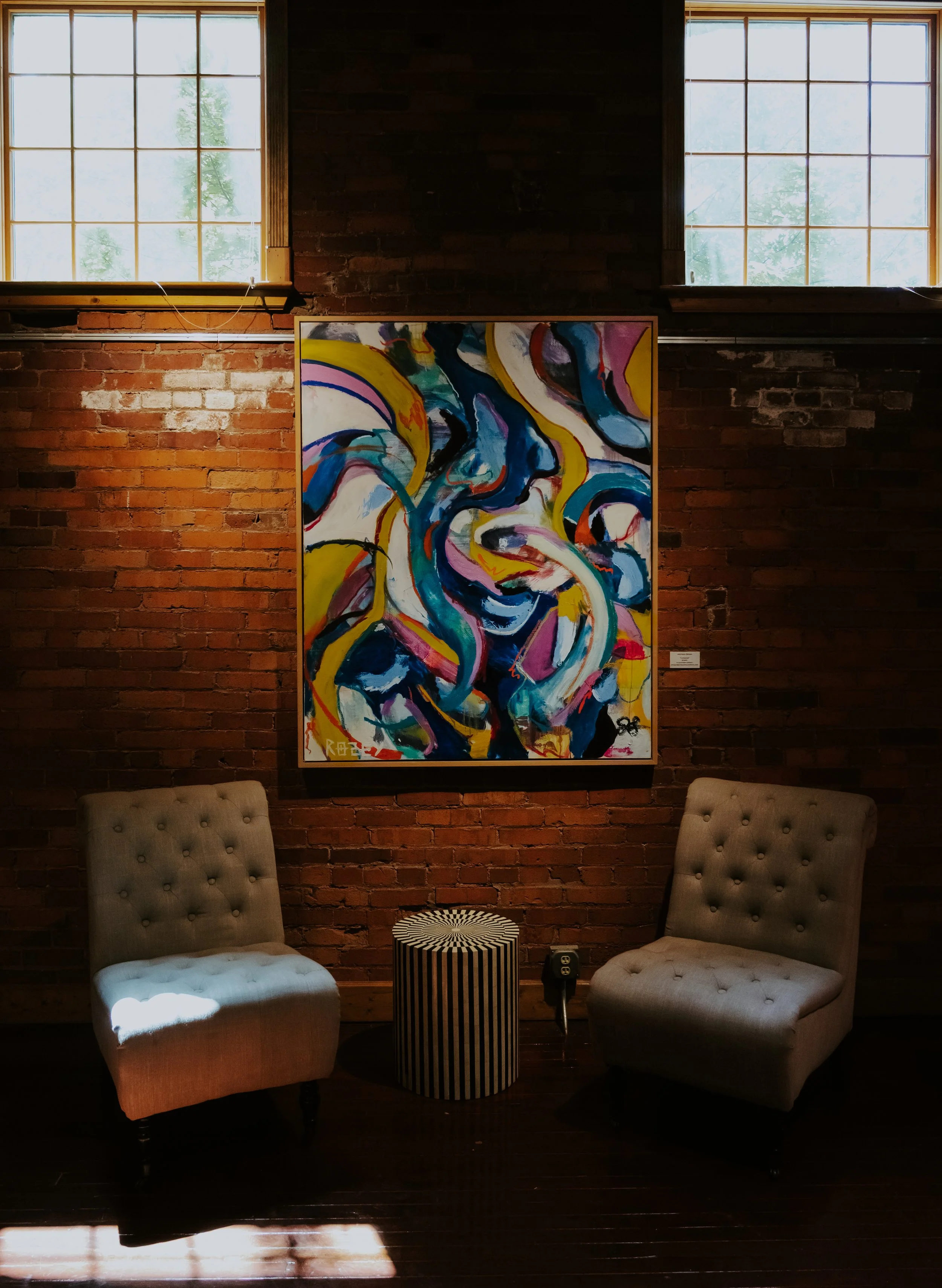 Interior of a room with a brick wall, two large windows, an abstract colorful painting, two beige tufted chairs, and a small black and white striped table.