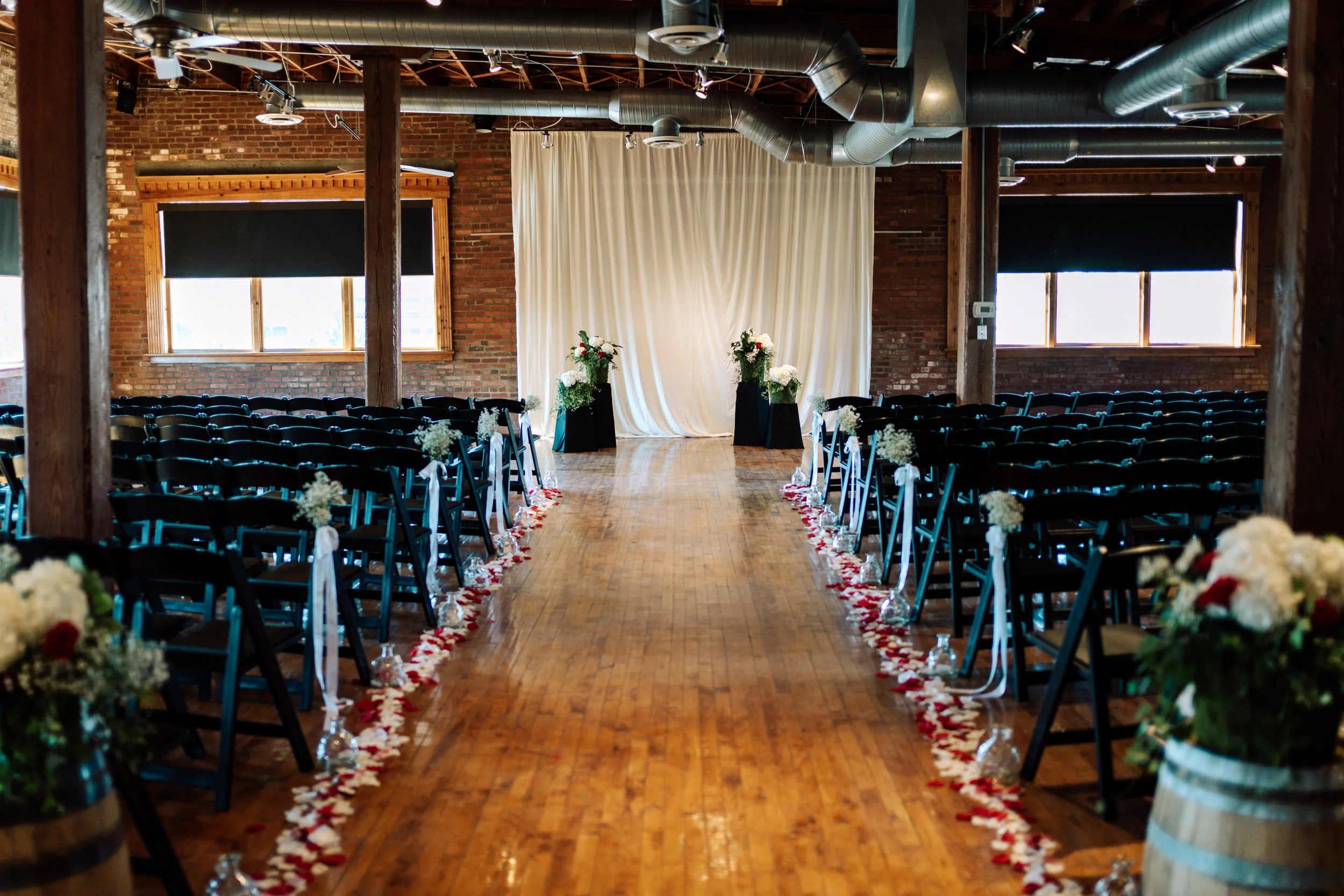 Indoor wedding ceremony setup with rows of black chairs decorated with white flowers along the aisle, floral arrangements in wooden barrels, a white backdrop curtain, and brick walls illuminated by windows.