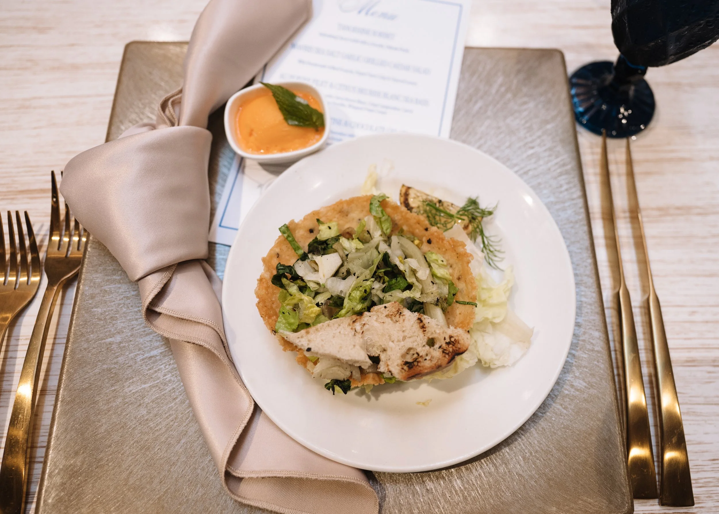 A plate with fried fish fillet topped with chopped lettuce, onions, and herbs, served with a lemon wedge. A small bowl of orange sauce with a green leaf is also on the table, along with a folded beige napkin, a fork, and a knife on a silver tray.