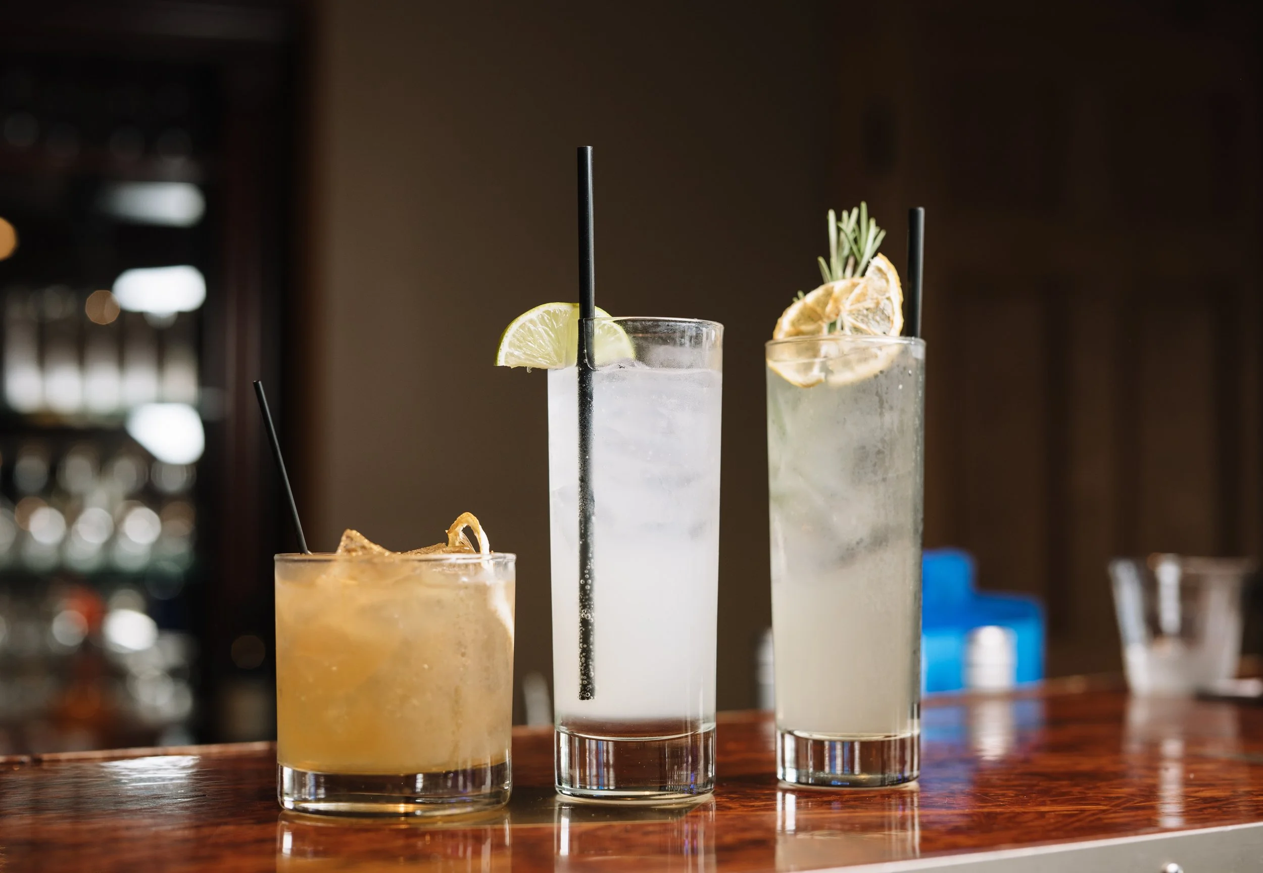 Three tall glasses with drinks garnished with lemon slices and herbs, one with a black straw, on a wooden bar counter.