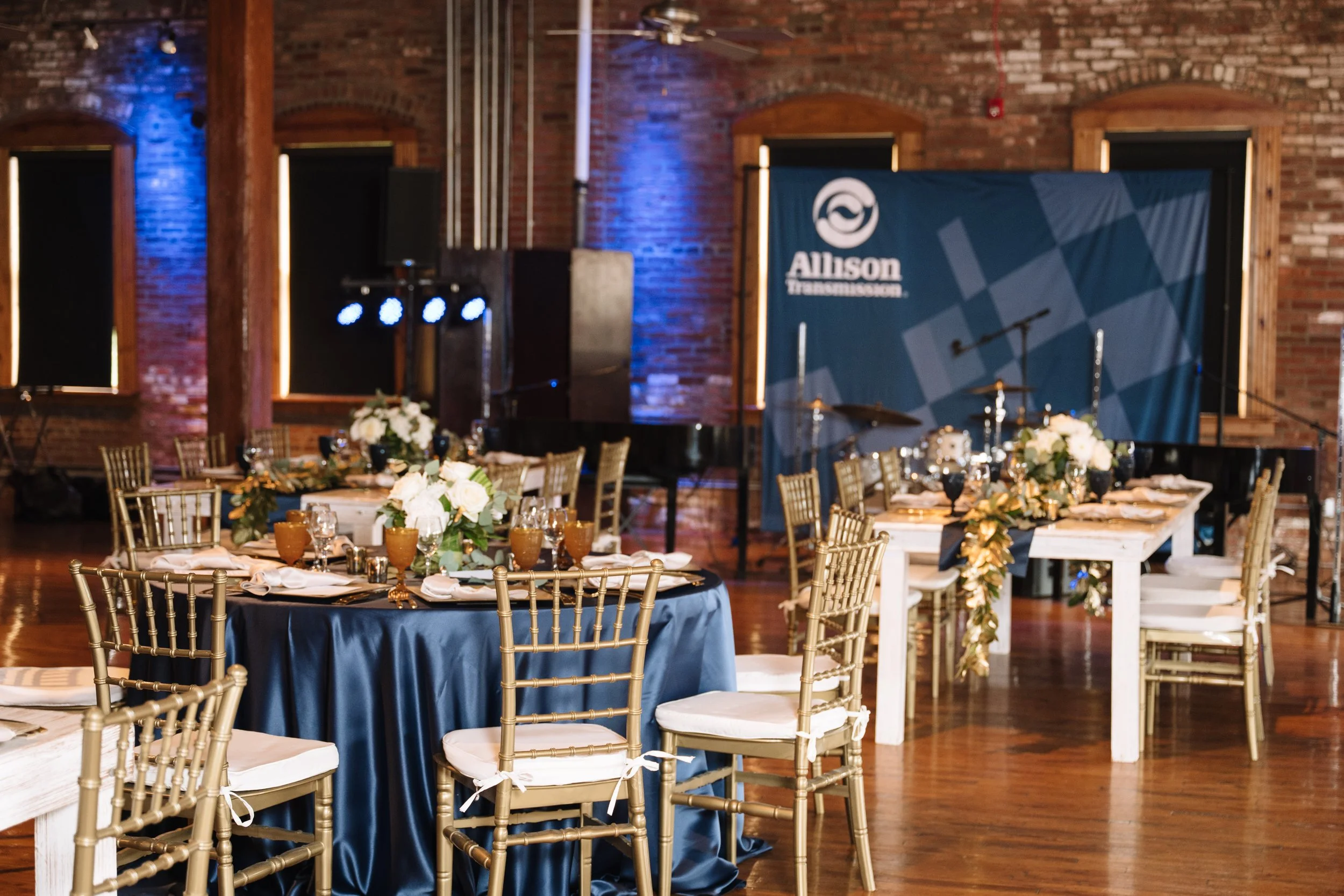 A decorated event venue with round tables covered in navy blue tablecloths, gold chairs, and floral centerpieces. There is a stage with a blue banner reading 'Allison Transformation' and musical instruments. The venue has exposed brick walls and wood