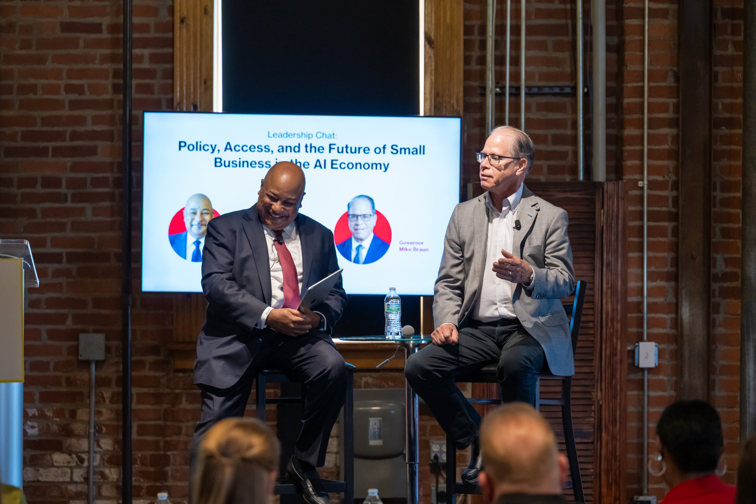 Two men, one bald and one with gray hair, sitting on stage in front of a screen. The screen displays the title "Policy, Access, and the Future of Small Business in the AI Economy" with pictures and names of speakers. The setting appears to be a confe