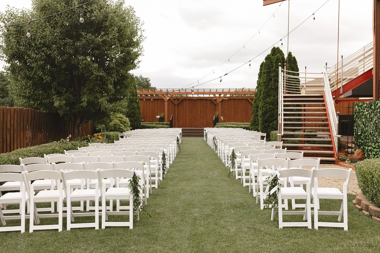 Outdoor wedding ceremony setup with white chairs decorated with greenery along a grassy aisle, facing a wooden stage. String lights hang overhead, and tall trees and a wooden fence surround the area.