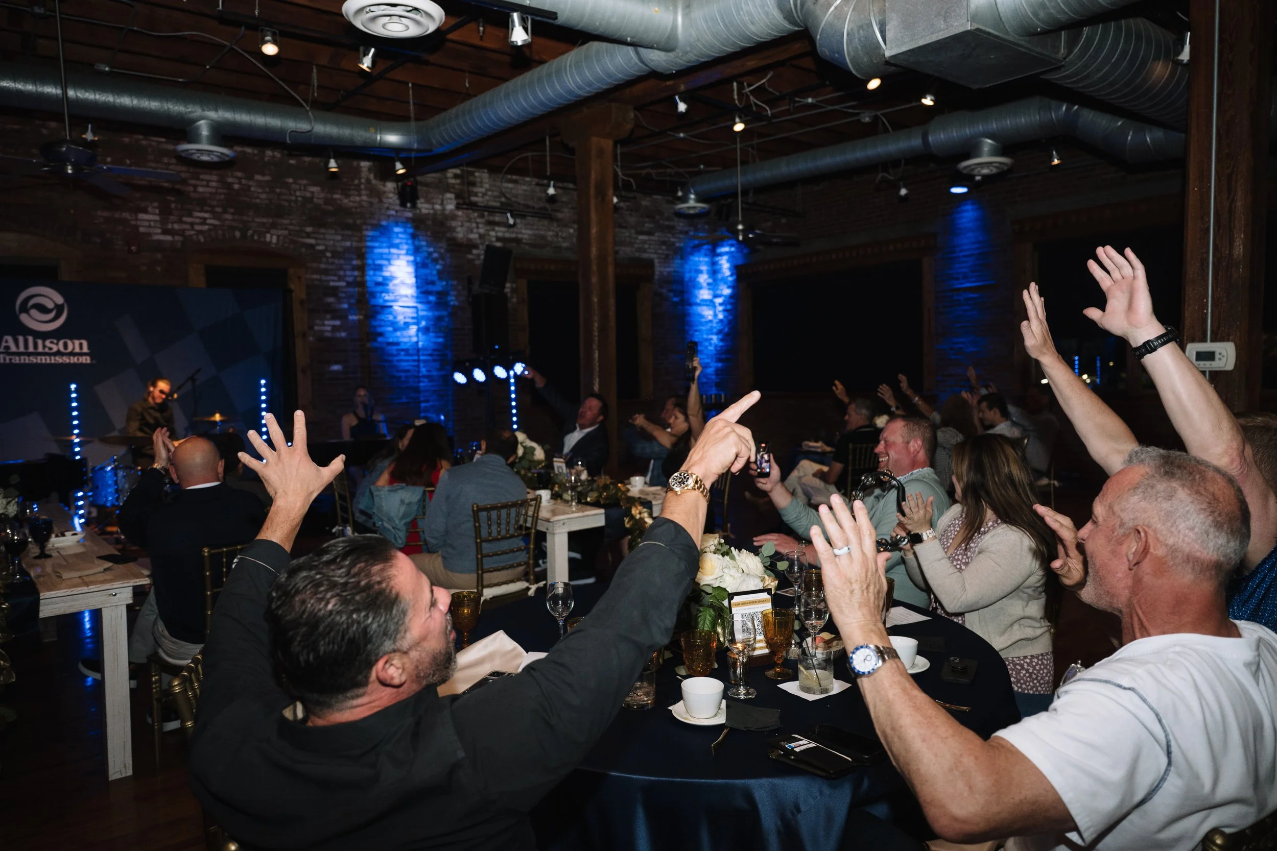 A large group of people seated at tables in a restaurant or banquet hall, raising their hands and engaging with the performers on stage. The background shows a stage with musicians, blue lighting, brick walls, and industrial ceiling pipes.