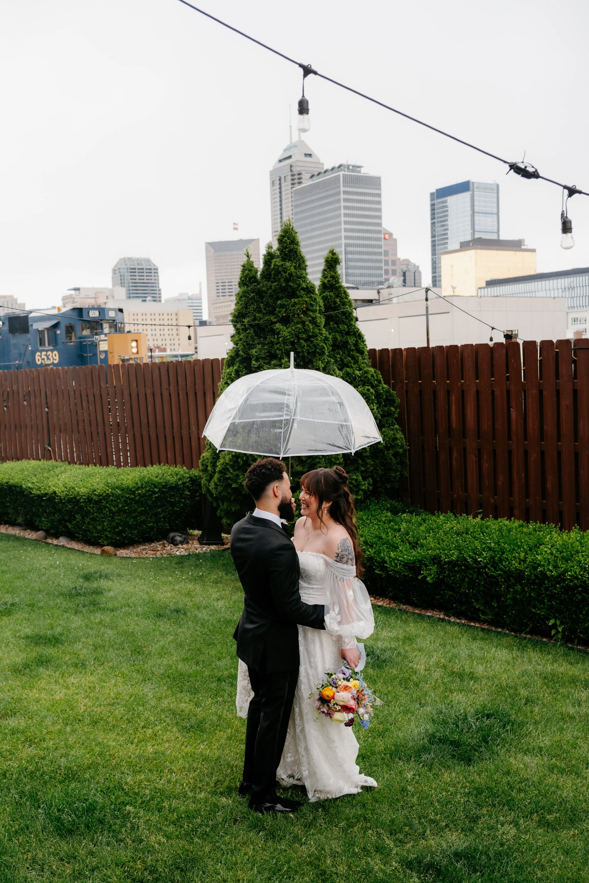 A bride and groom standing in a grassy area with a city skyline in the background. They are holding hands, looking at each other, with a clear umbrella and a bouquet of flowers. The bride is wearing a white wedding dress, and the groom is in a black 