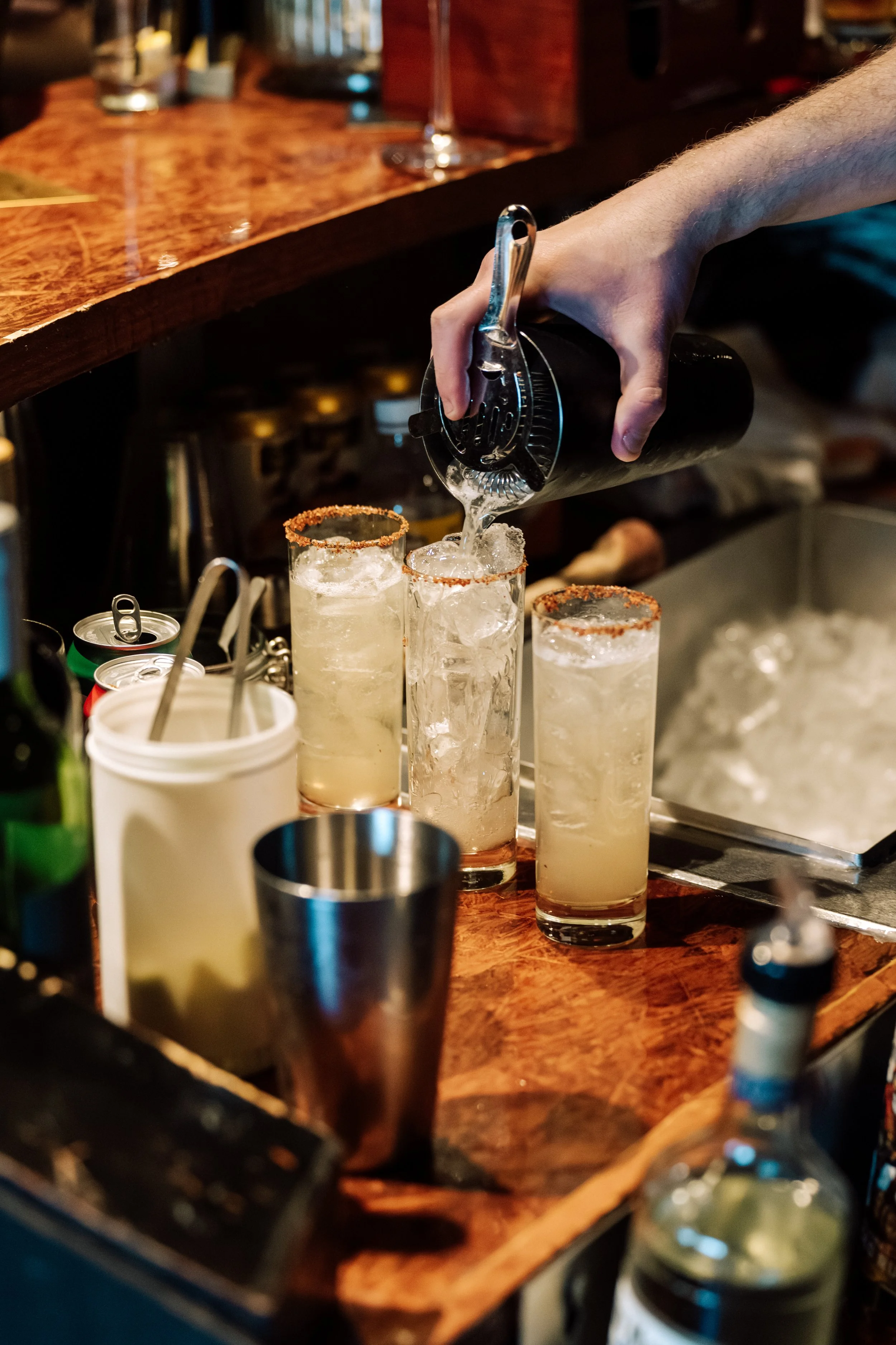 A person is pouring liquid into tall glasses with salted rims, likely making cocktails, at a bar with a wooden counter.
