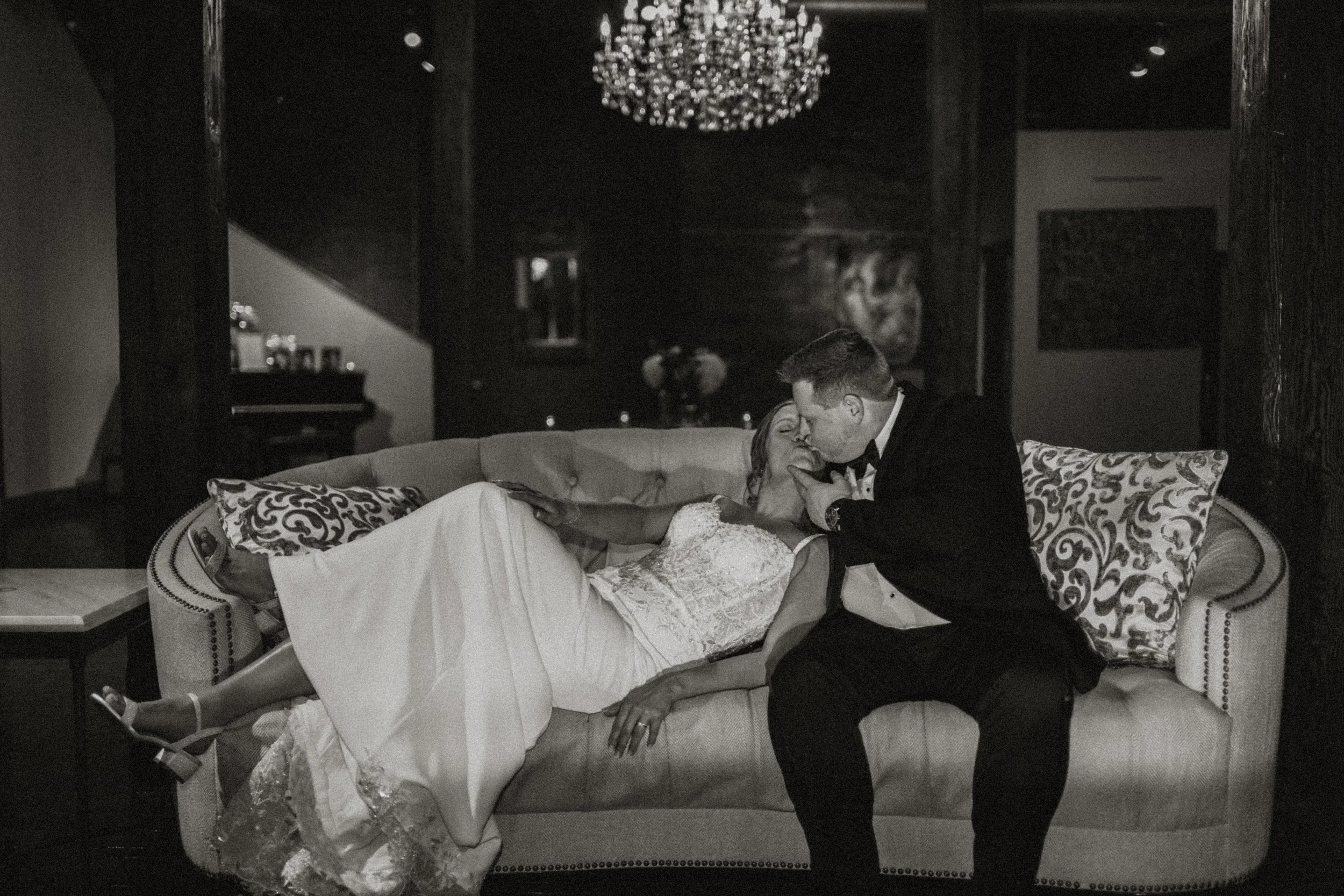 A bride and groom share a kiss, with the bride lying on a sofa and the groom leaning over her, in a dimly lit room with elegant decor, including a chandelier and framed art on the walls.