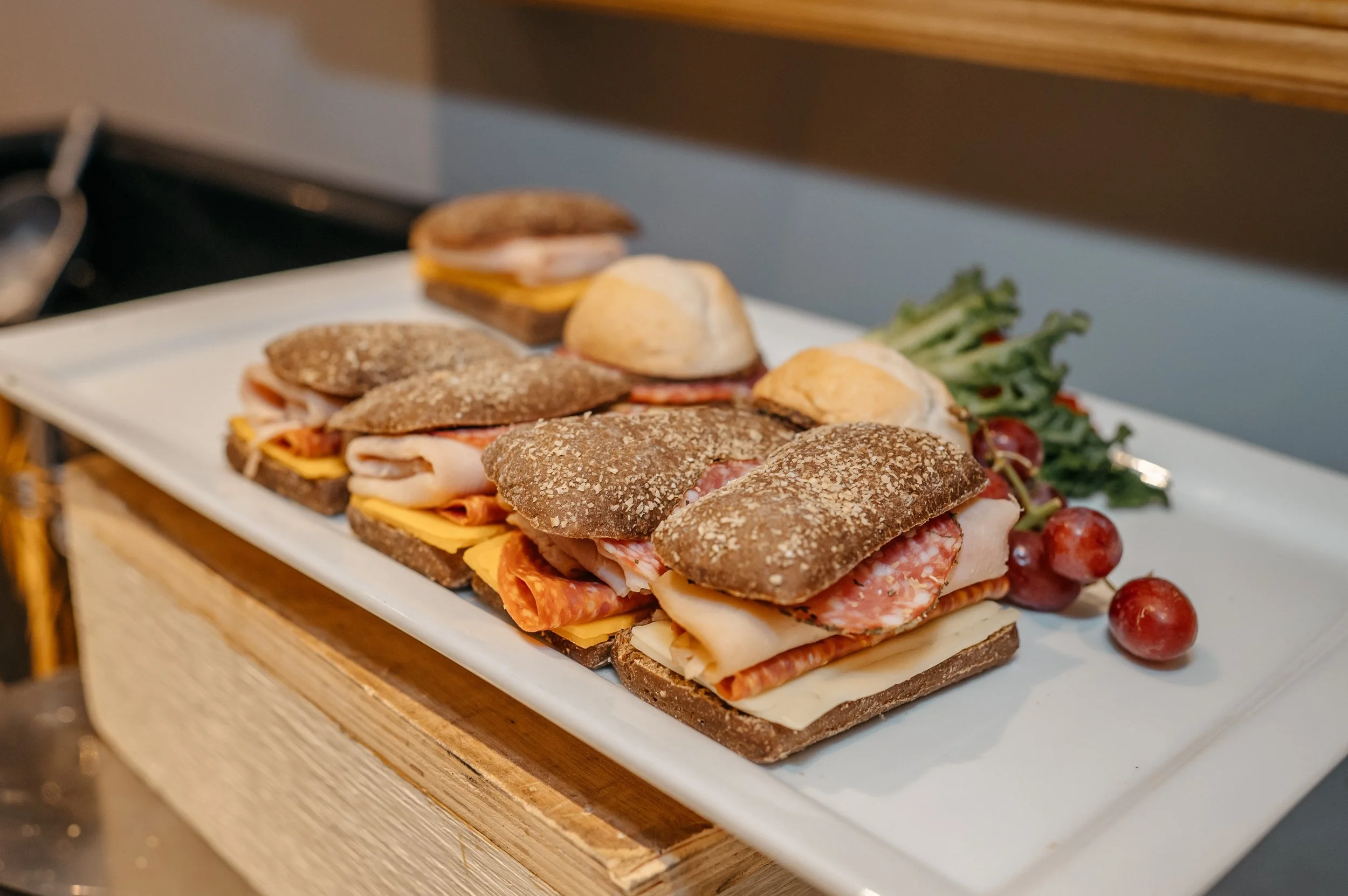 Assorted deli sandwiches arranged on a rectangular white plate with greens and red grapes on the side, placed on a wooden surface.