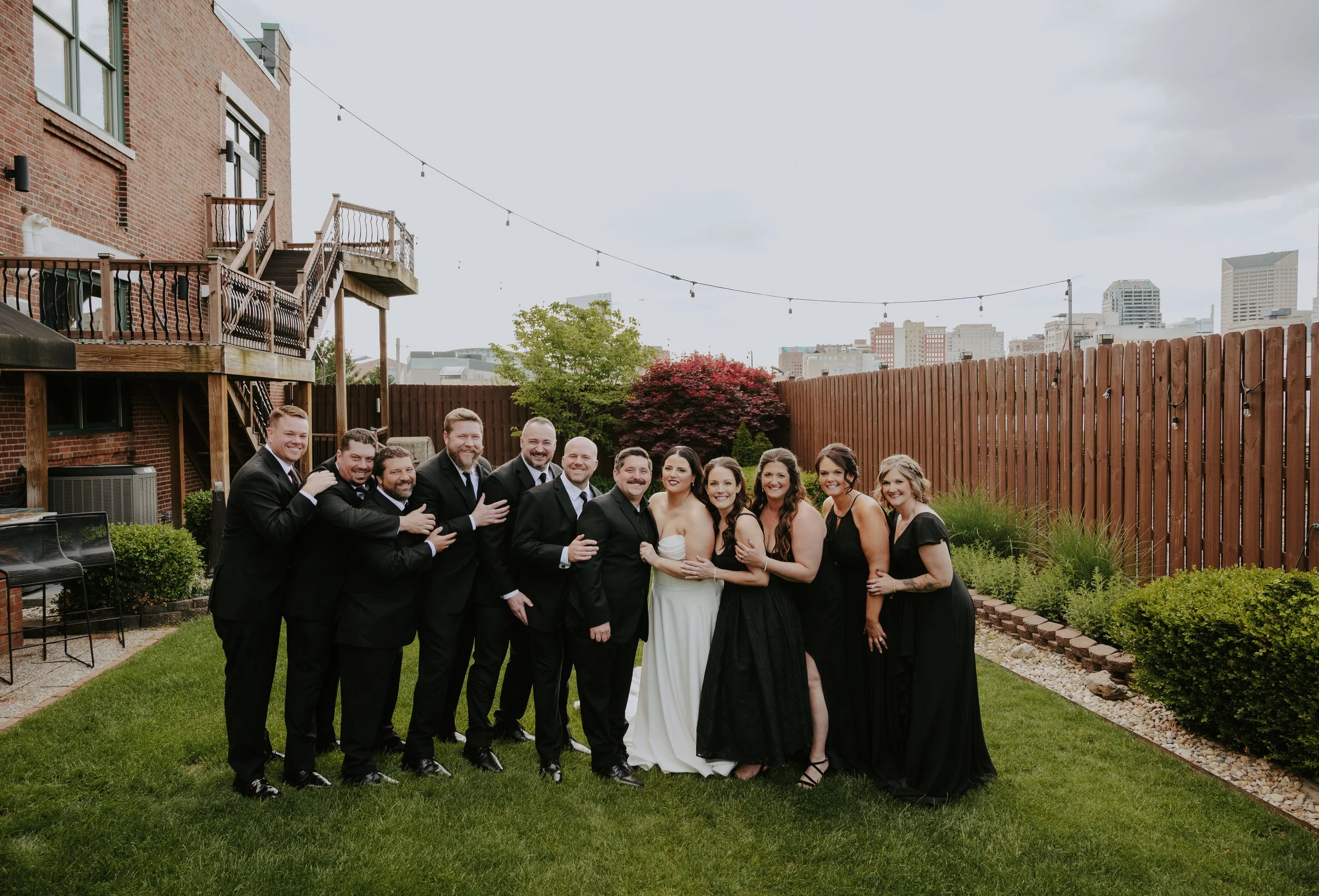 A group of people in formal attire posing in a backyard with a wooden fence, trees, and an urban city skyline in the background on a cloudy day.