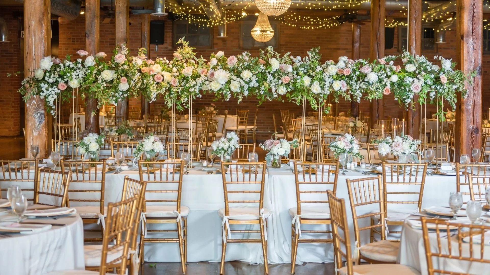 Long banquet table set with white tablecloths, glassware, and elegant floral centerpieces. Overhead, a large floral arrangement of white and blush roses with greenery hangs above the table. The background features warm brick walls, wooden beams, stri