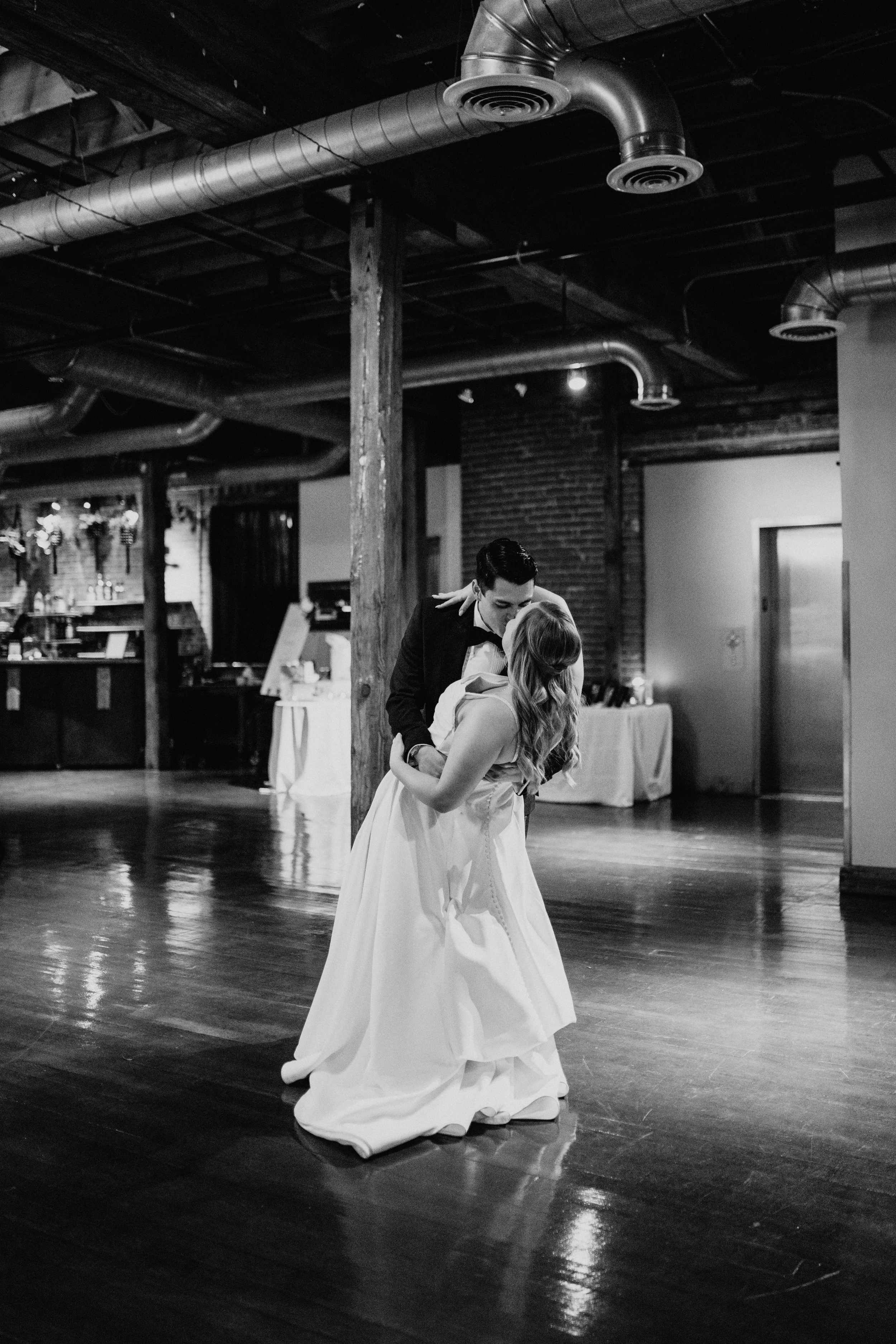 A bride and groom share a dance in a rustic indoor venue with exposed brick walls, wooden beams, and ventilation ducts.