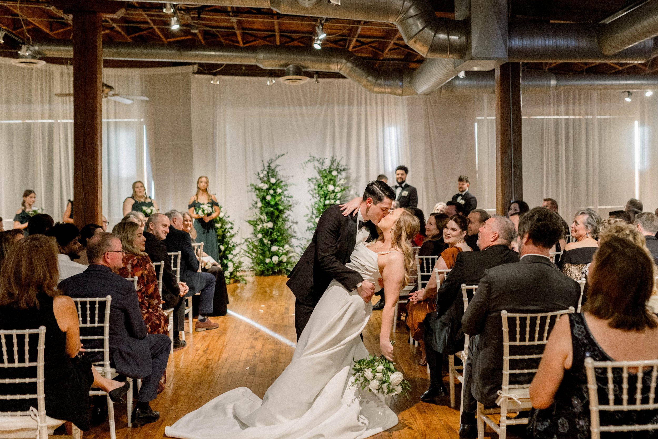 A wedding ceremony with the bride and groom sharing a kiss, surrounded by seated guests in an indoor venue with wooden beams and industrial ductwork. The bride is in a white gown, and the groom is in a black tuxedo.
