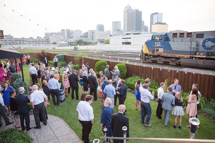 People attending a social gathering outdoors near a train passing on elevated tracks in an urban area with buildings in the background.