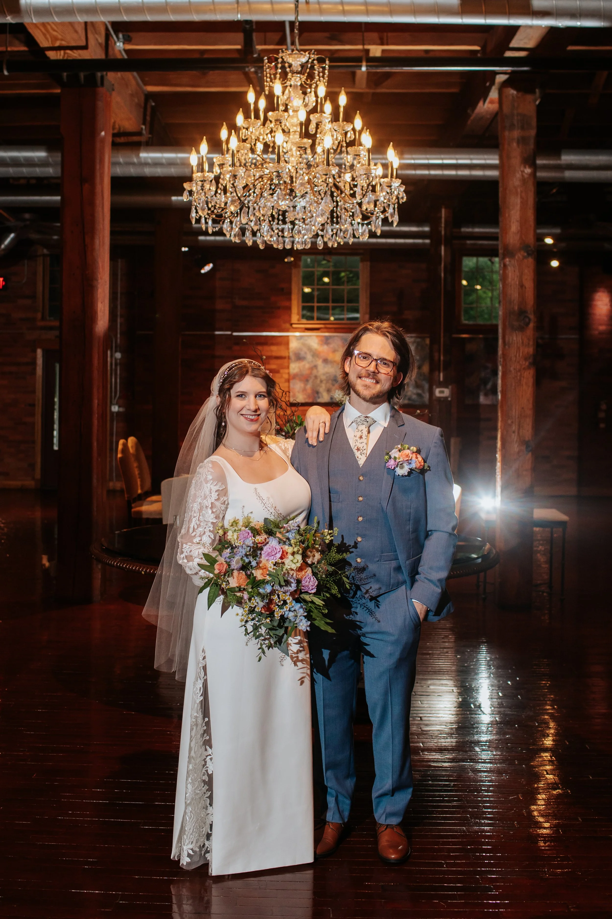 A newlywed couple standing in a warmly lit wooden hall with a large crystal chandelier overhead. The bride wears a white lace wedding dress with long sleeves and a veil, holding a colorful bouquet of flowers. The groom in a gray suit with a floral ti