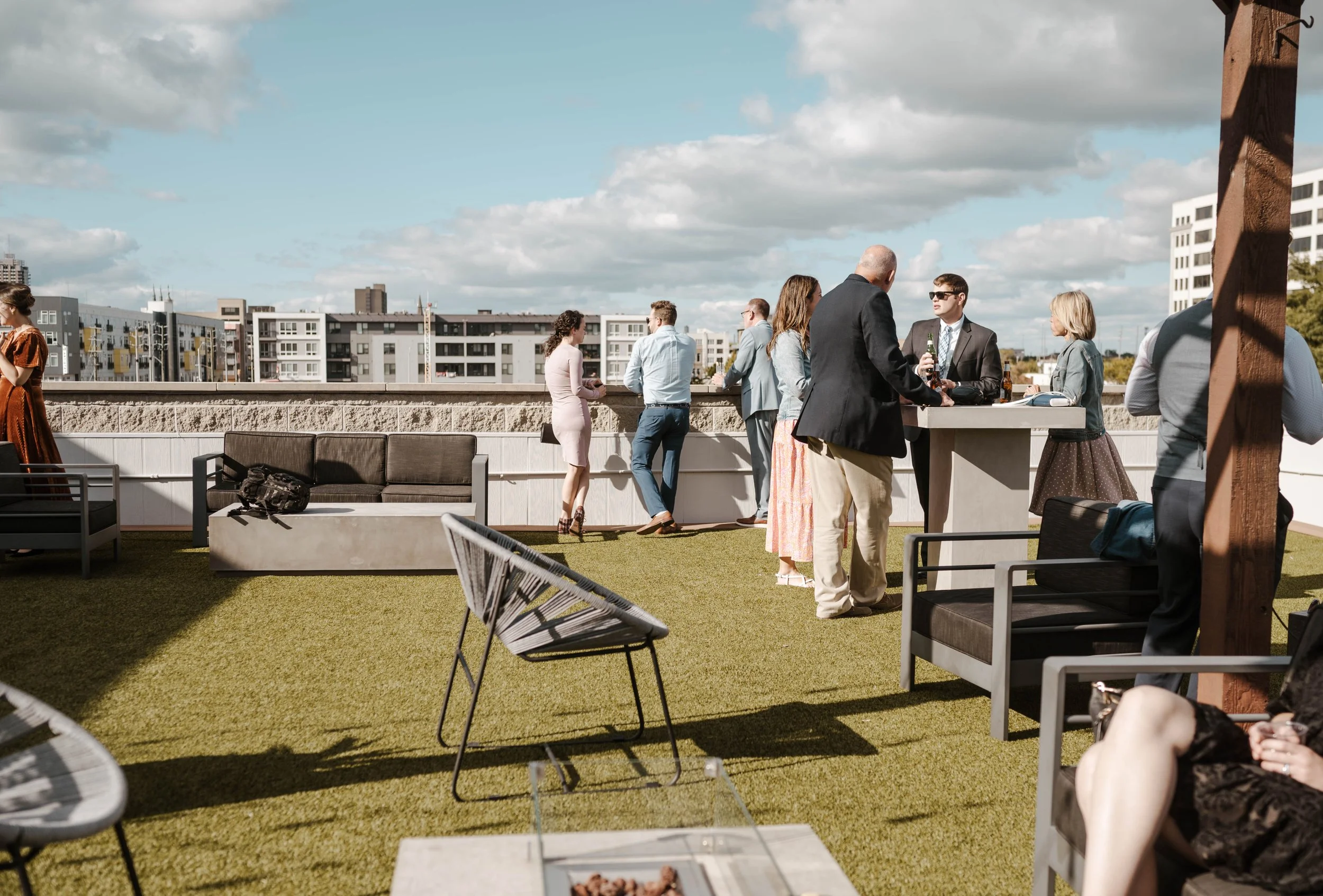 People socializing on a rooftop patio with outdoor seating, tables, and cityscape in the background on a partly cloudy day.