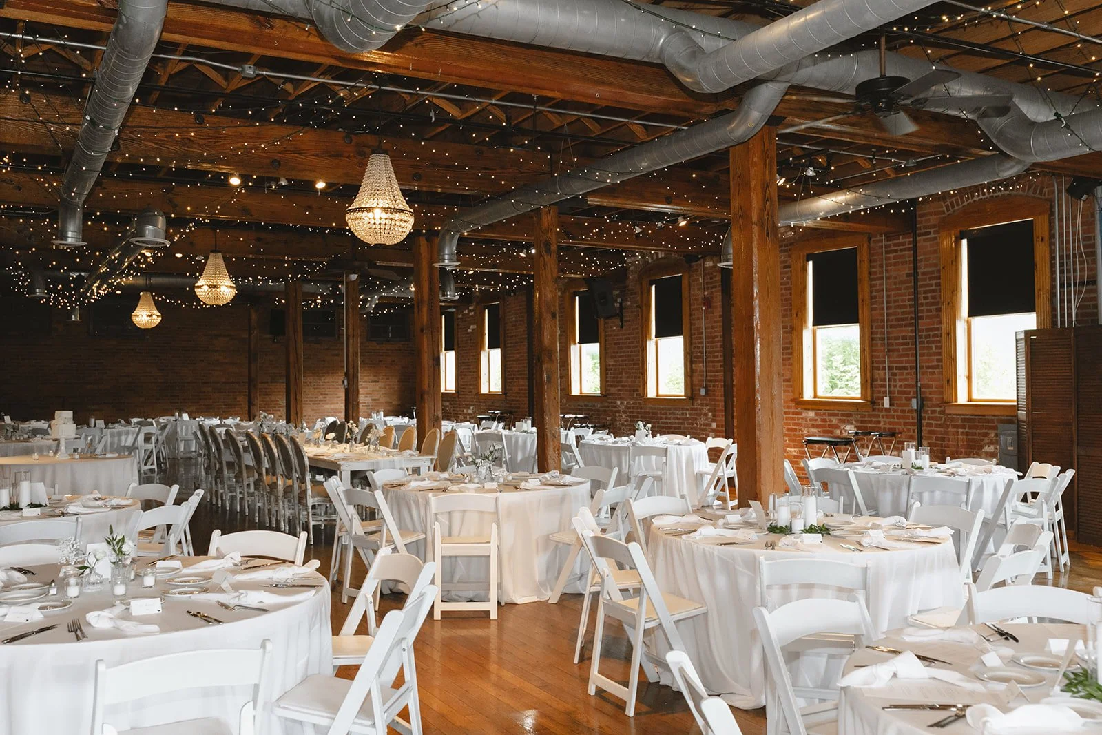 A spacious banquet hall with round tables covered in white tablecloths, set for a formal event with plates, silverware, and candles. The hall has wooden beams, brick walls, large windows with black blinds, and is decorated with string lights and chan