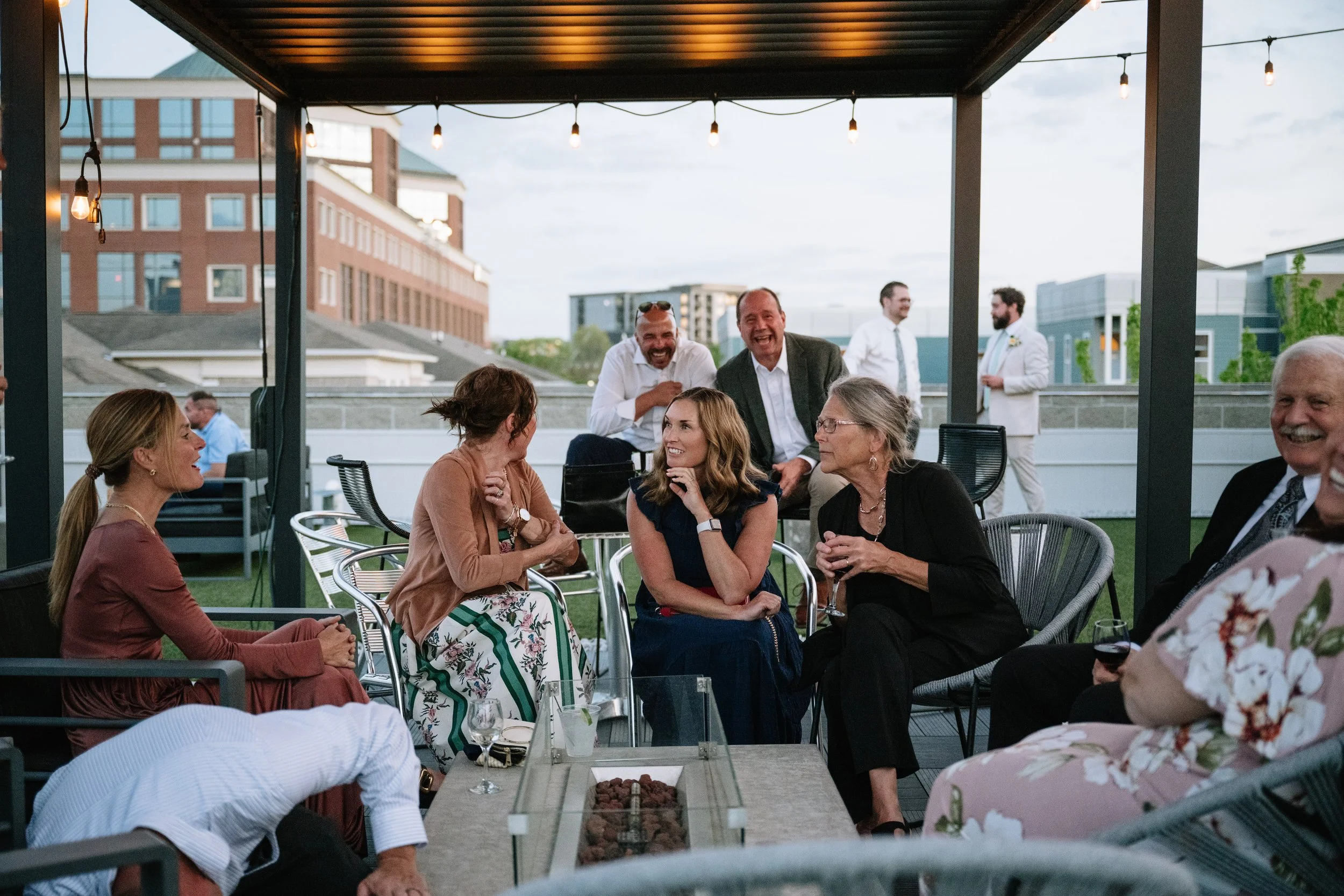 Group of people enjoying a social gathering outdoors on a covered rooftop patio, with some standing and others sitting, engaging in conversations and laughter, during the evening with decorative string lights.