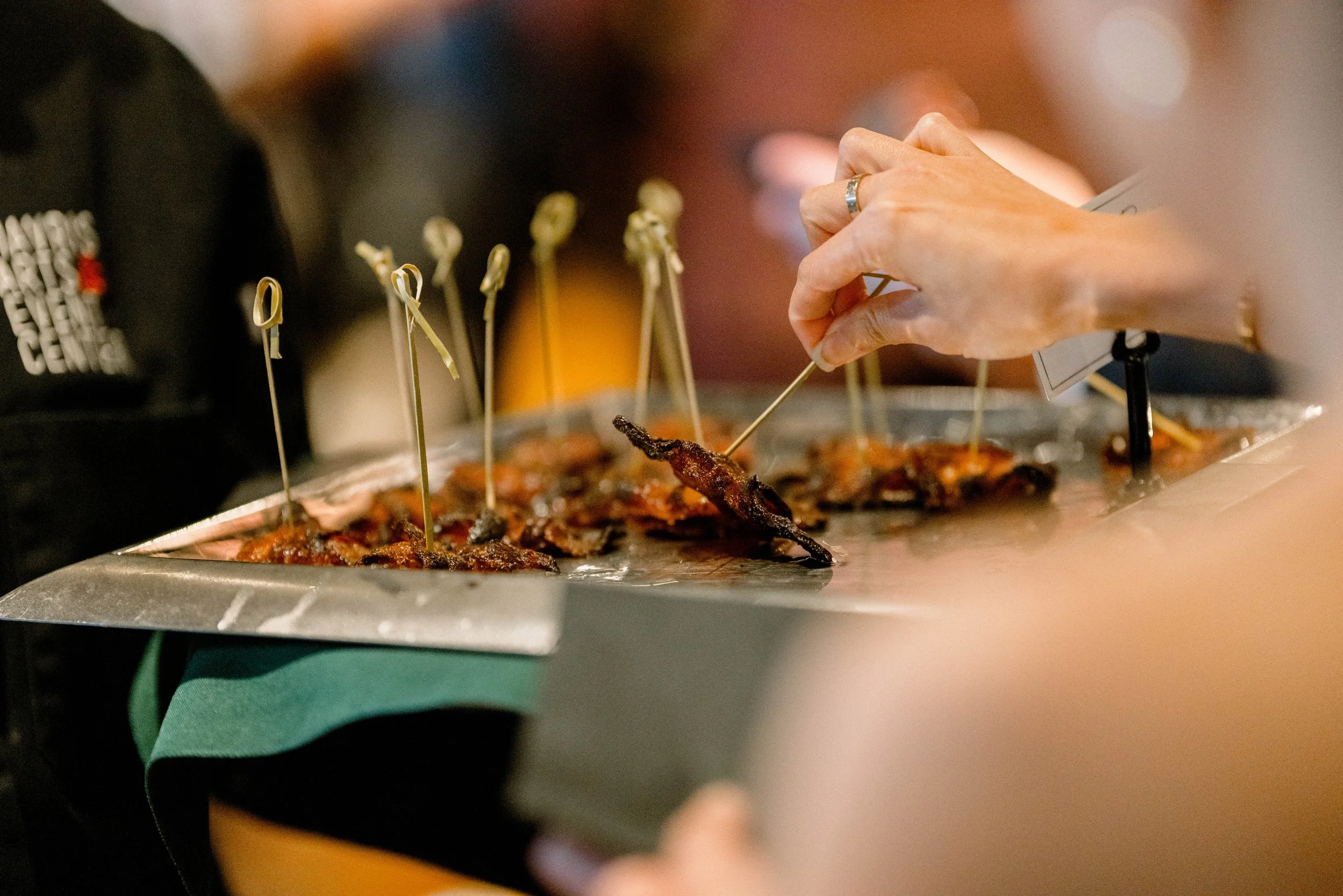 A hand picks up skewers of grilled meat or kebabs from a serving tray at a restaurant or buffet.