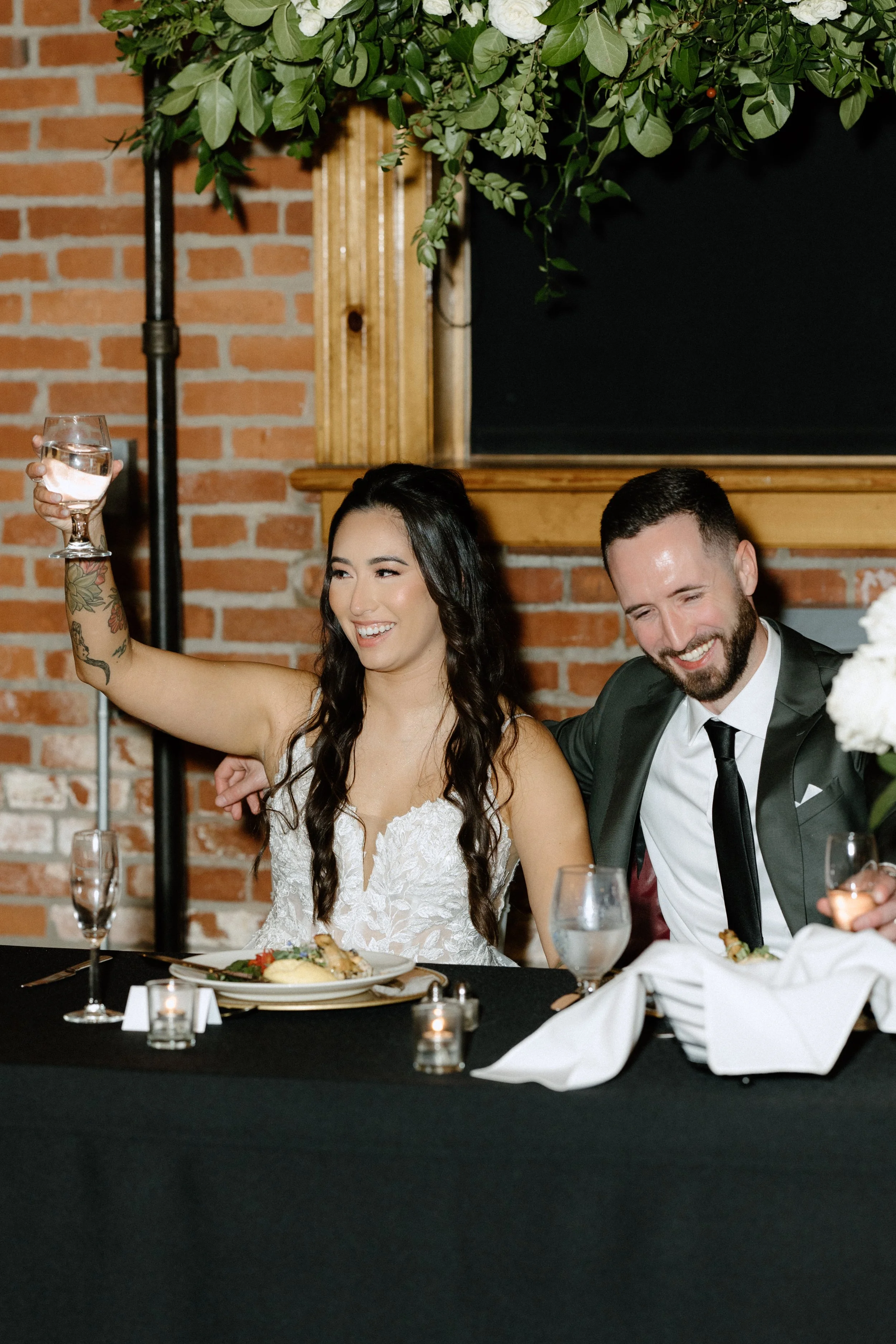 A woman with long dark hair, wearing a white lace dress, is raising a glass of water and smiling. A man with a beard, wearing a dark suit and tie, is sitting next to her, also smiling. They are at a formal event with a brick wall in the background, a