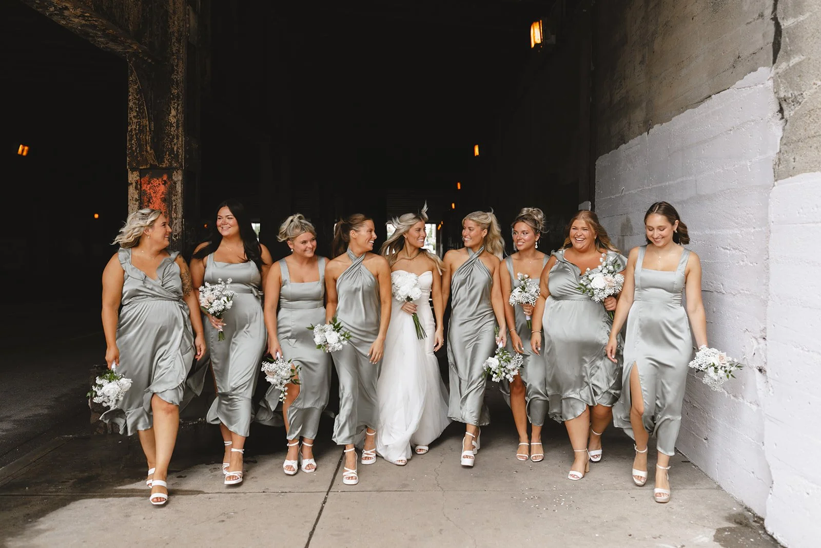A bride and her bridesmaids walk together in an urban setting, all dressed in matching gray satin dresses, holding bouquets of white flowers, and smiling.
