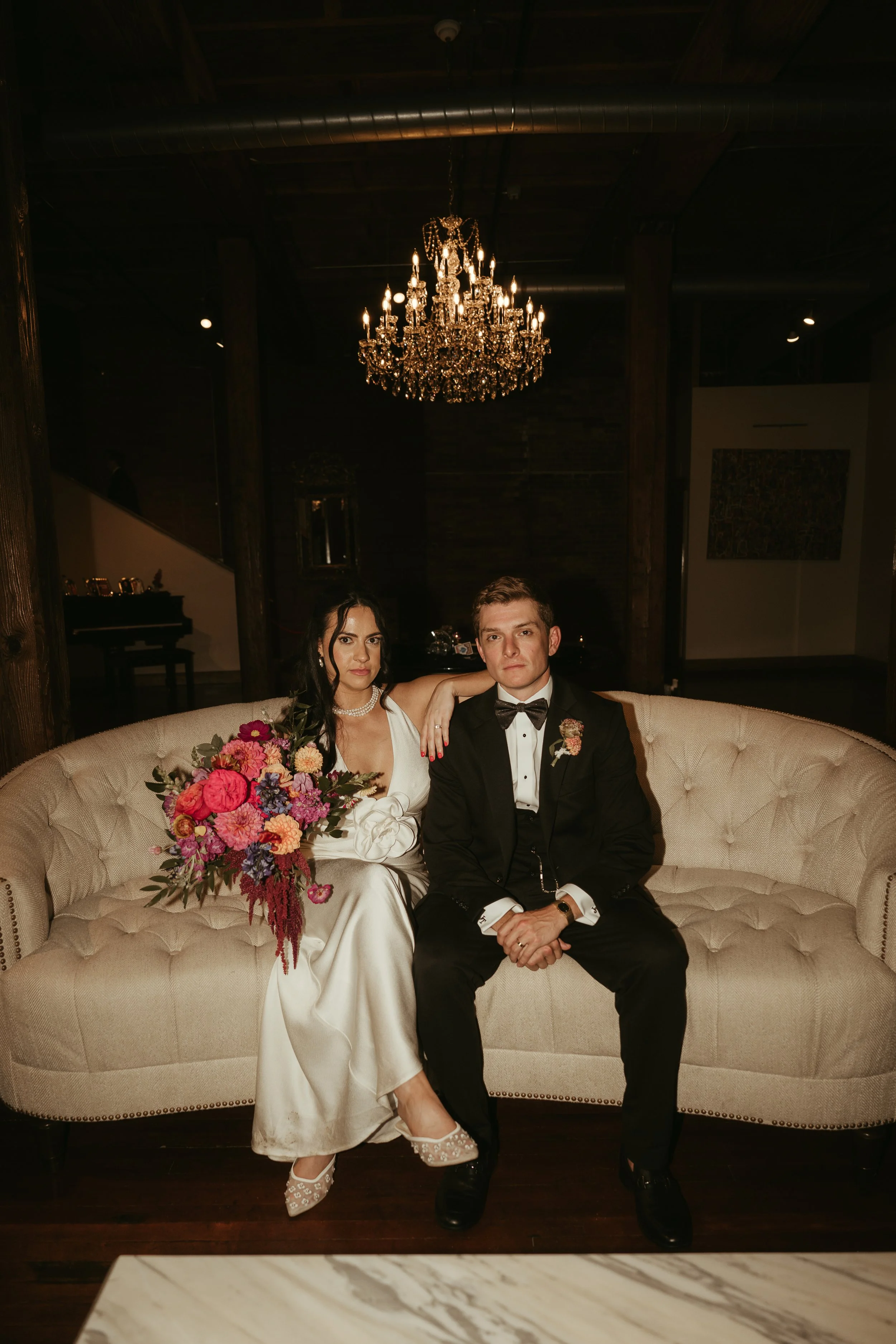 A bride and groom sitting on a beige tufted sofa in an elegant, dimly lit room with a large crystal chandelier overhead. The bride is wearing a white wedding gown, holding a colorful bouquet, and the groom is dressed in a black tuxedo with a bow tie,