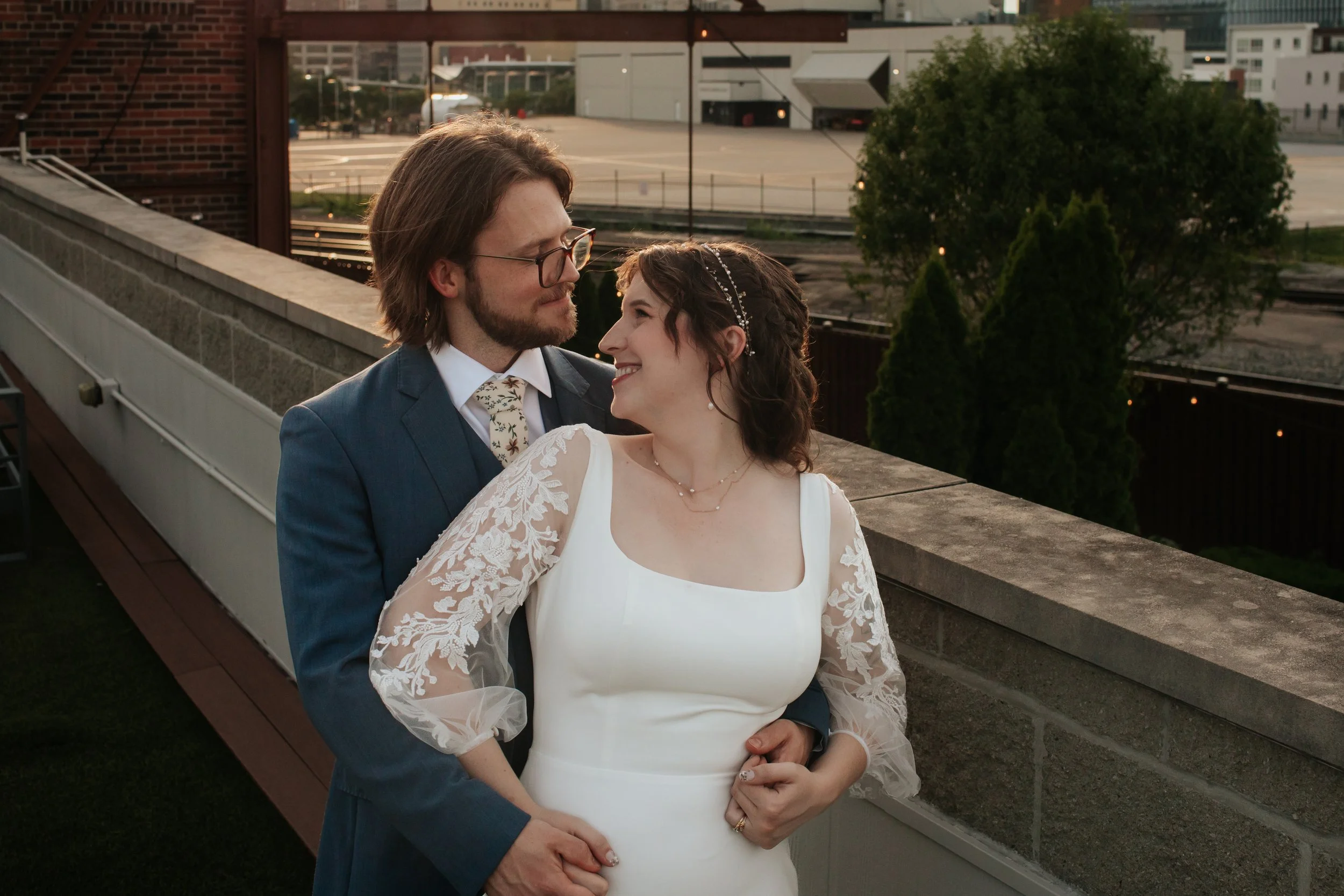 A bride and groom smiling and looking into each other's eyes on a rooftop overlooking a city. The bride is wearing a white wedding dress with lace sleeves and a pearl headband. The groom is dressed in a dark suit, white shirt, and patterned tie, wear