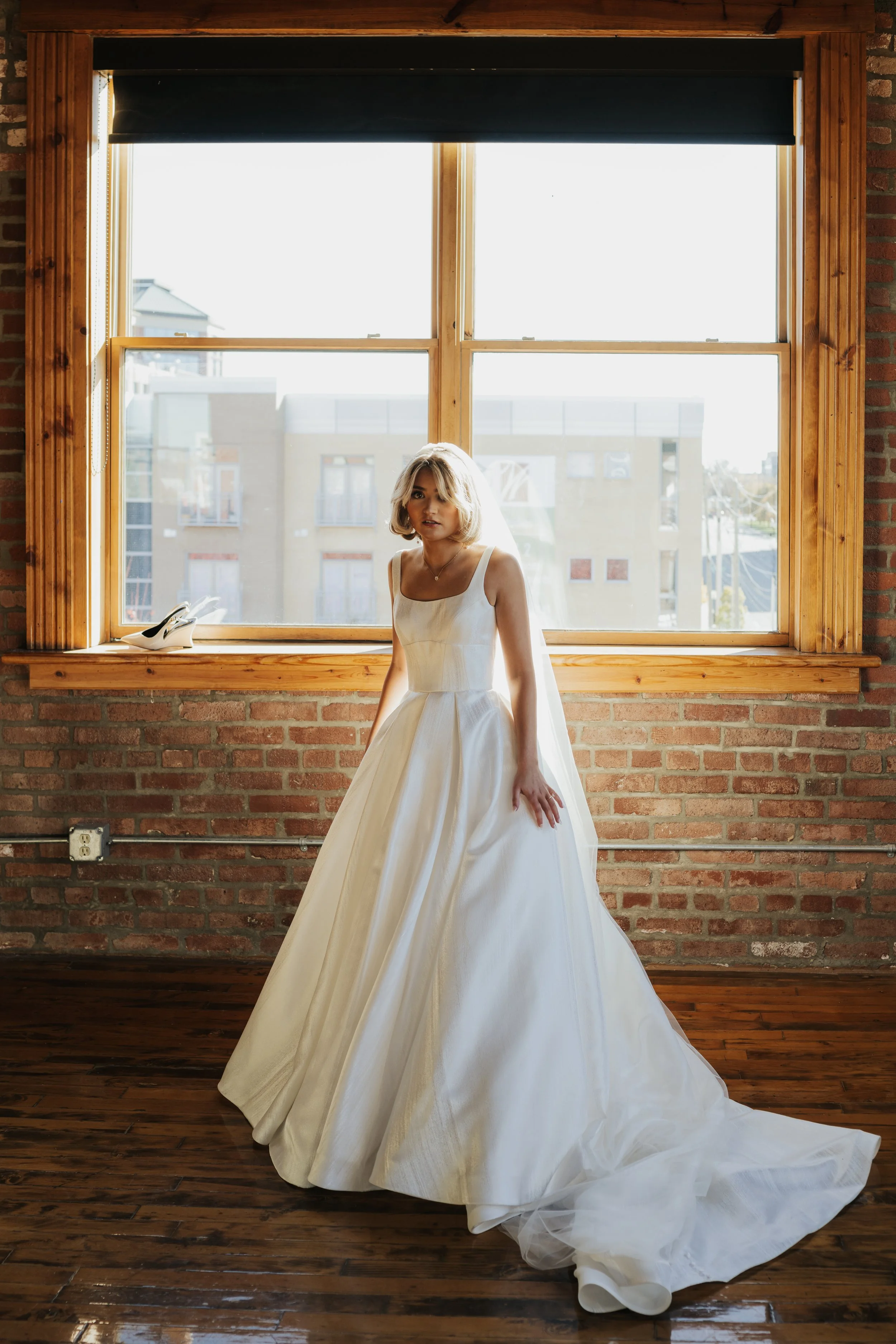 A woman in a wedding dress standing in front of a large window with a brick wall behind her, a pair of white shoes on the window sill, natural light illuminating her and the dress.