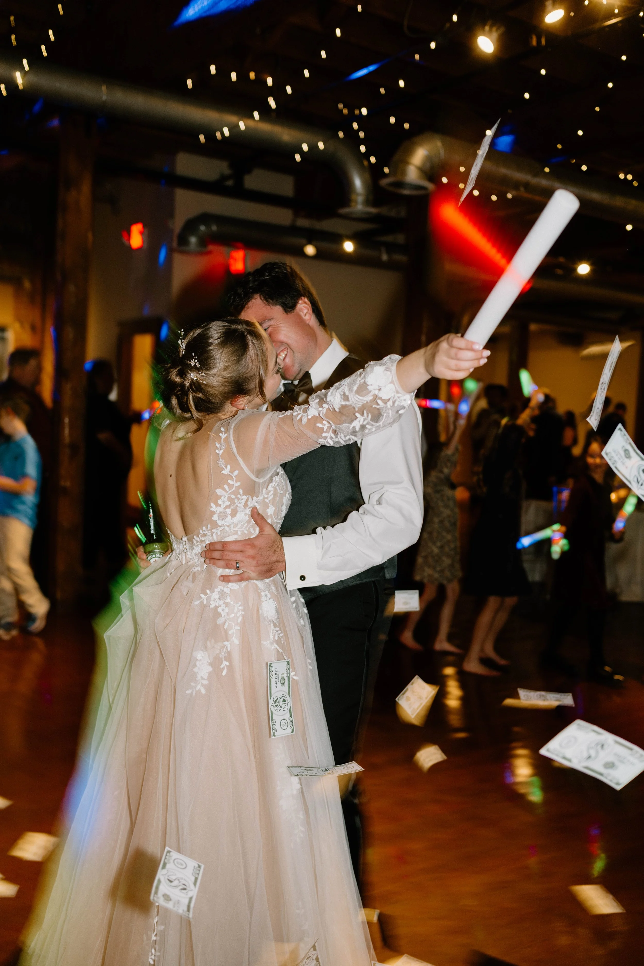 A bride and groom dancing, holding small rolled papers and surrounded by falling money, at a wedding reception indoors with string lights and other guests in the background.
