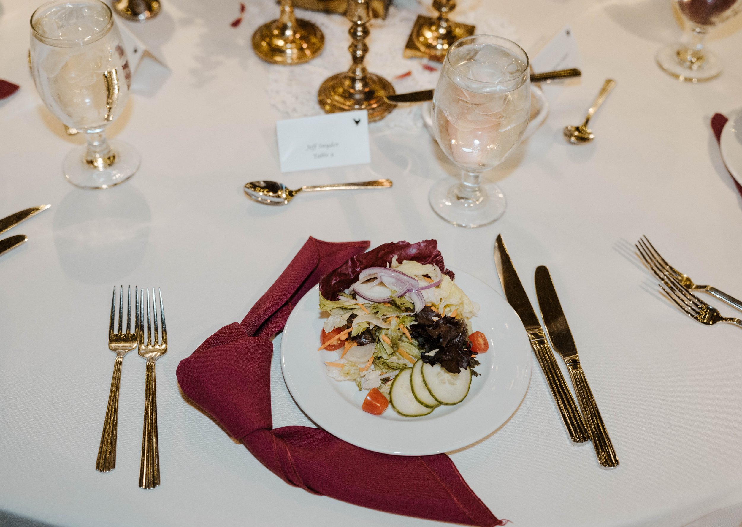 A dining table set for a meal, with gold utensils and glasses of water with ice. There is a salad on a white plate with sliced cucumbers, cherry tomatoes, and lettuce. A burgundy napkin is placed underneath the plate.