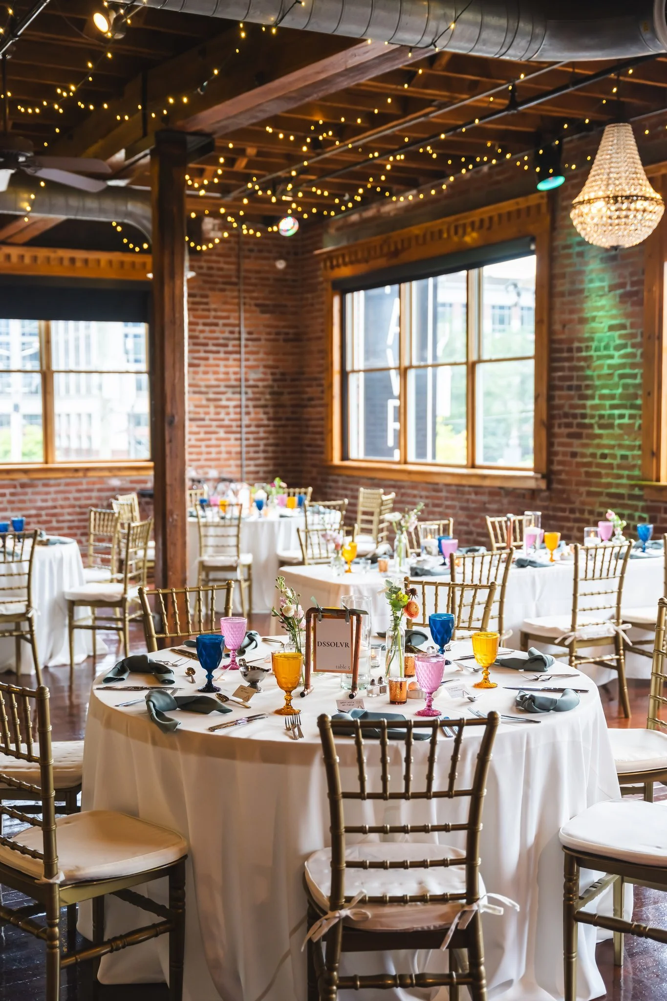 Indoor wedding reception setup with round tables covered with white tablecloths, colorful glassware, floral centerpieces, and gold chairs in a rustic brick-walled venue decorated with string lights and a chandelier.